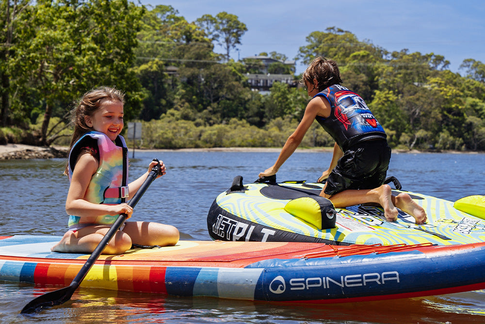 Two kids in Jetpilot youth life jackets paddling a SUP, showing safe gear for groms on the water