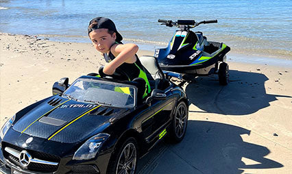 Young kid on a ride-on toy car towing a miniature Jetpilot electric jet ski trailer along a sandy beach
