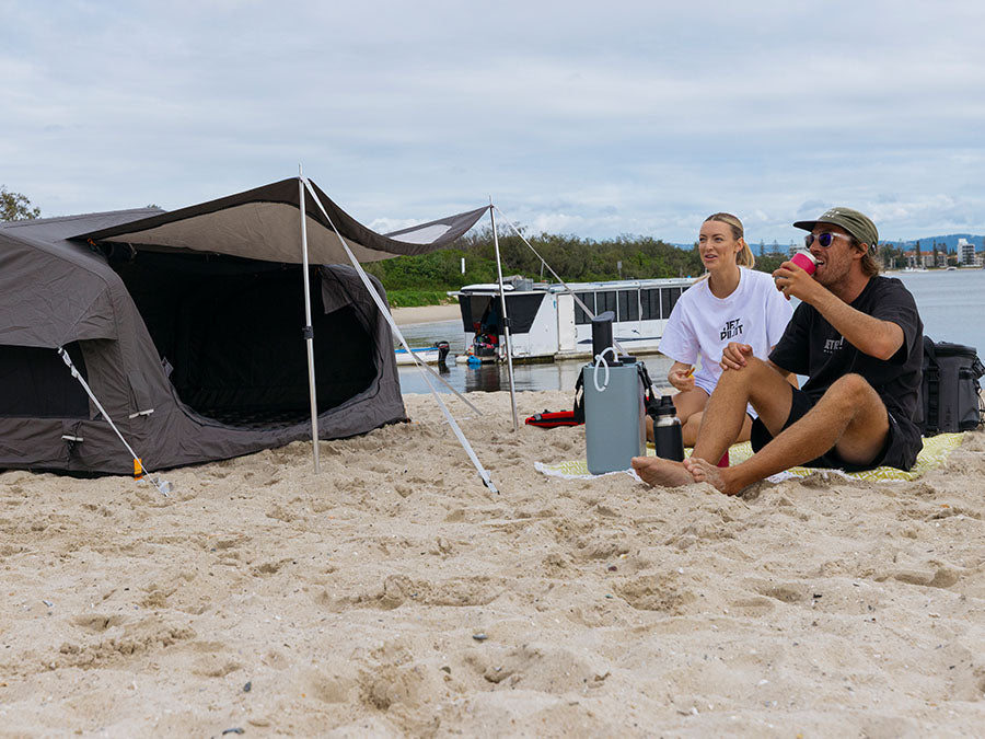 Couple beach camping with a swag tent, cooler and jet ski gear set up on the sand, showing key beach camping essentials