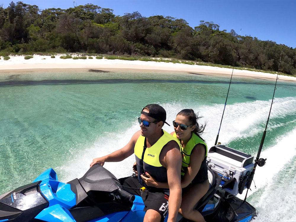 Couple in hi-vis Jetpilot PFDs cruising a fishing-rigged jet ski past a secluded sandy beach