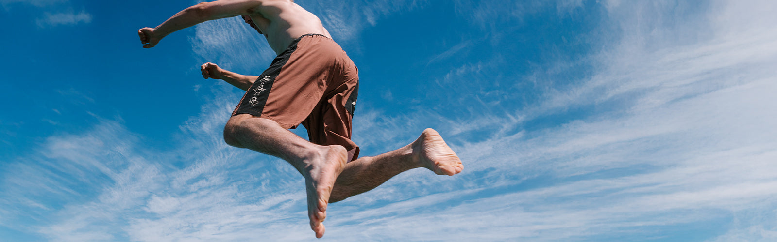Bloke mid-jump against a blue summer sky wearing brown Jetpilot mens board shorts, barefoot