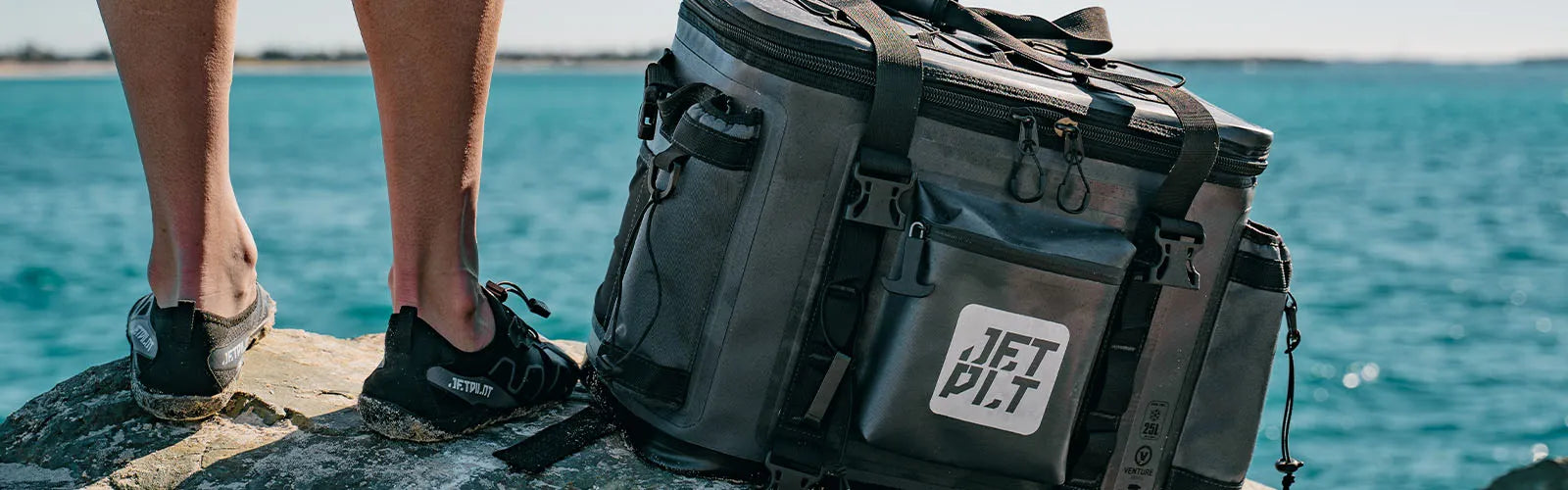 Jetpilot Venture cooler bag and jetski accessories on rocks beside turquoise water, feet in water shoes