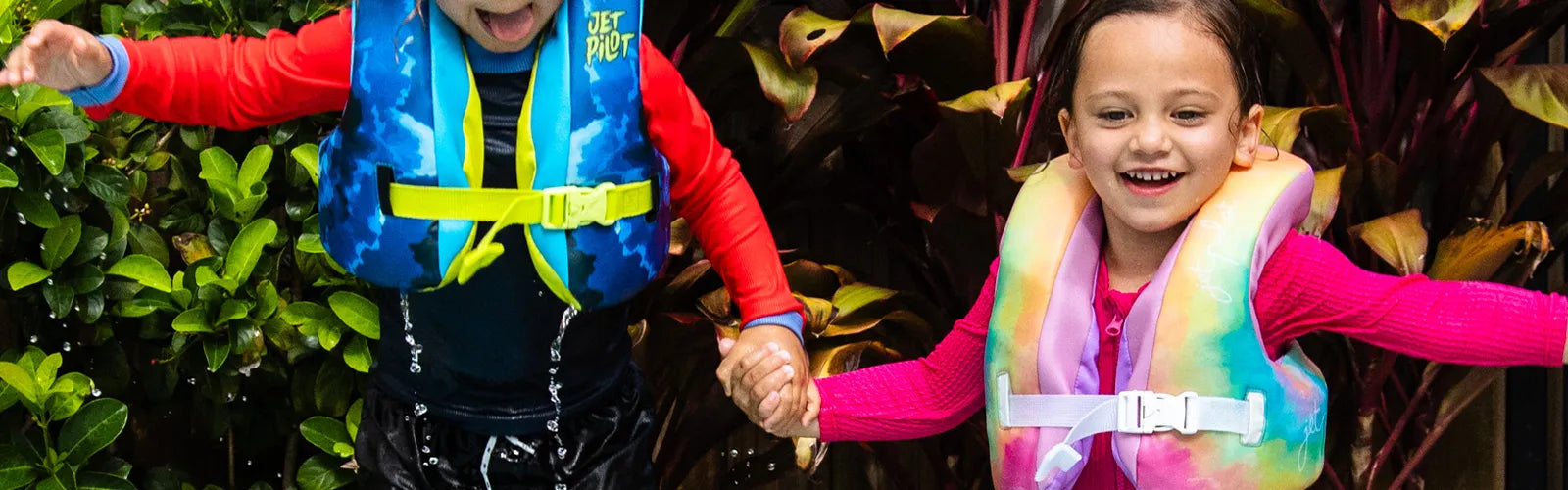Two kids smiling in Jetpilot kids life jackets, a blue cloud print and a rainbow pastel vest