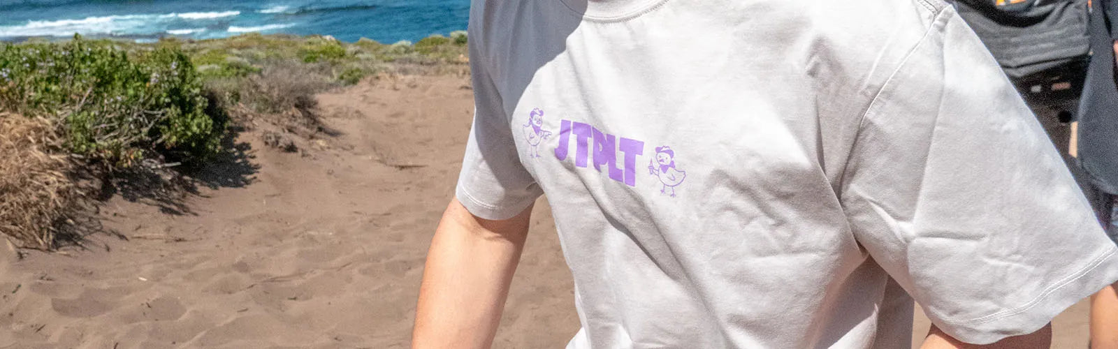 Back view of a man in a grey Jetpilot JTPLT print tee walking the sand dunes above a surf beach