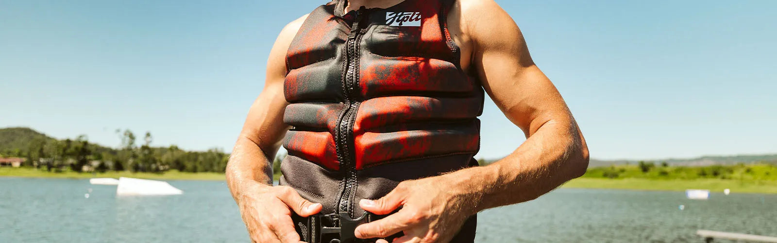 Bloke fastening a red and black Jetpilot mens life jacket by the lake at a wake park