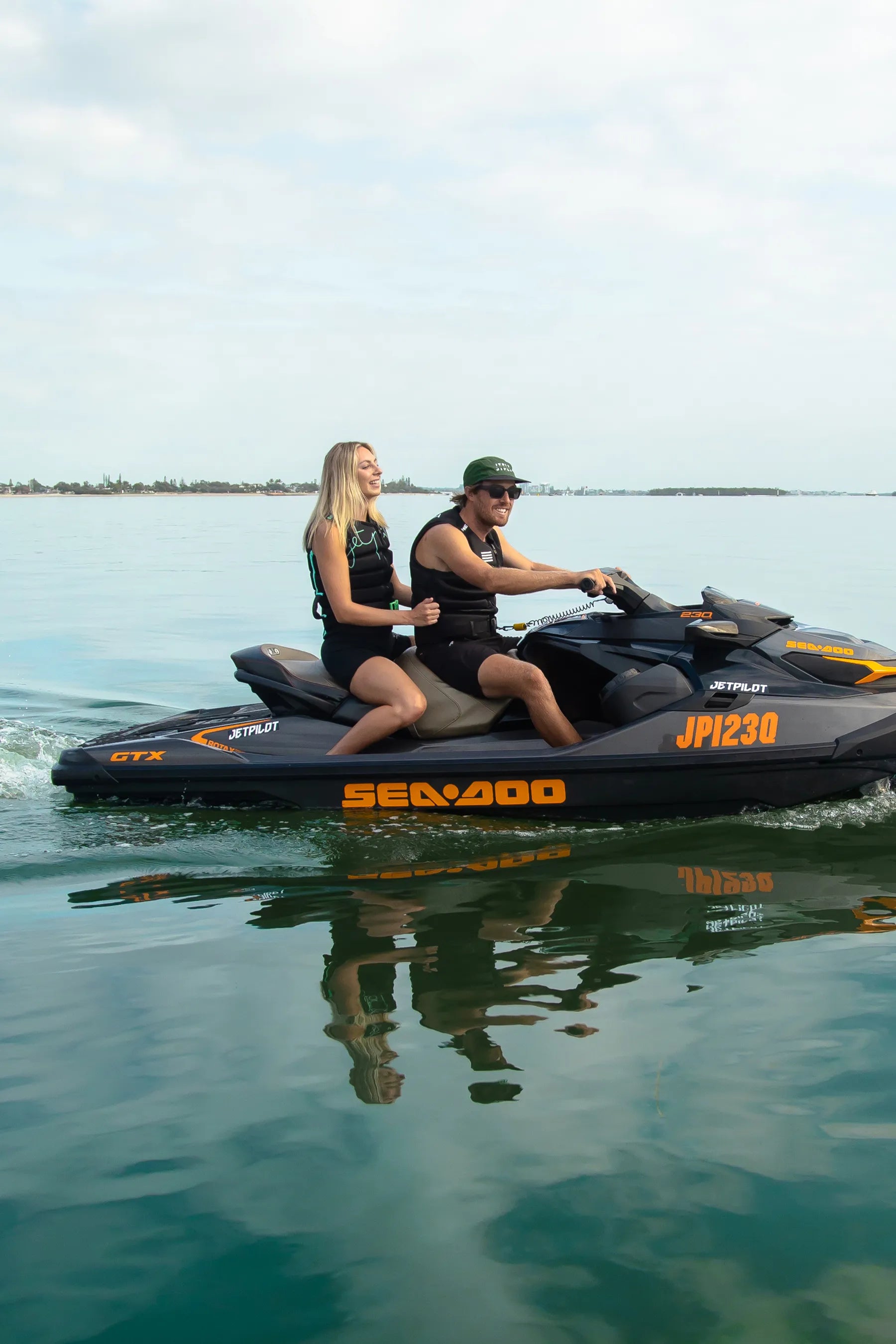 Two people riding a Sea-Doo jet ski, in Jetpilot Life Jackets on calm water with a clear sky.
