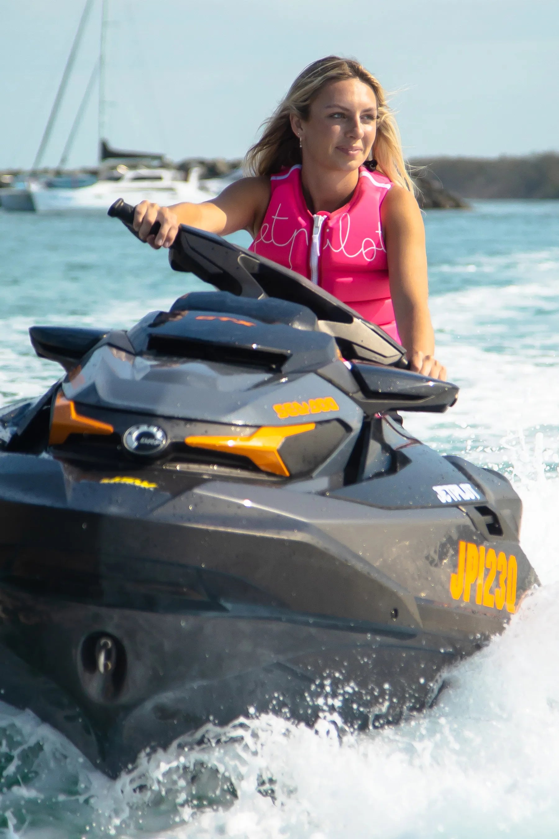Woman riding a Sea-Doo watercraft, in a Jetpilot life jacket on a sunny day with boats in the background.
