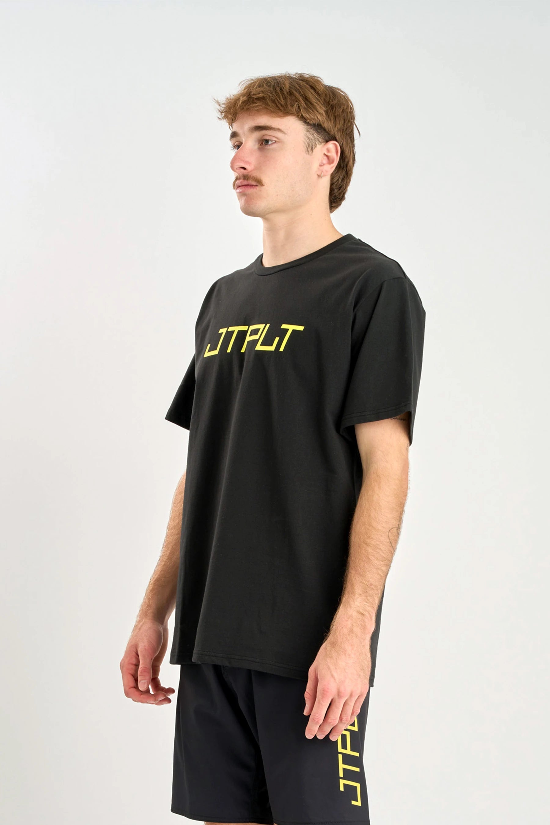 Person wearing a Jetpilot Vault SS Tee - Black t-shirt with yellow text and black shorts with yellow text on a white backgrou