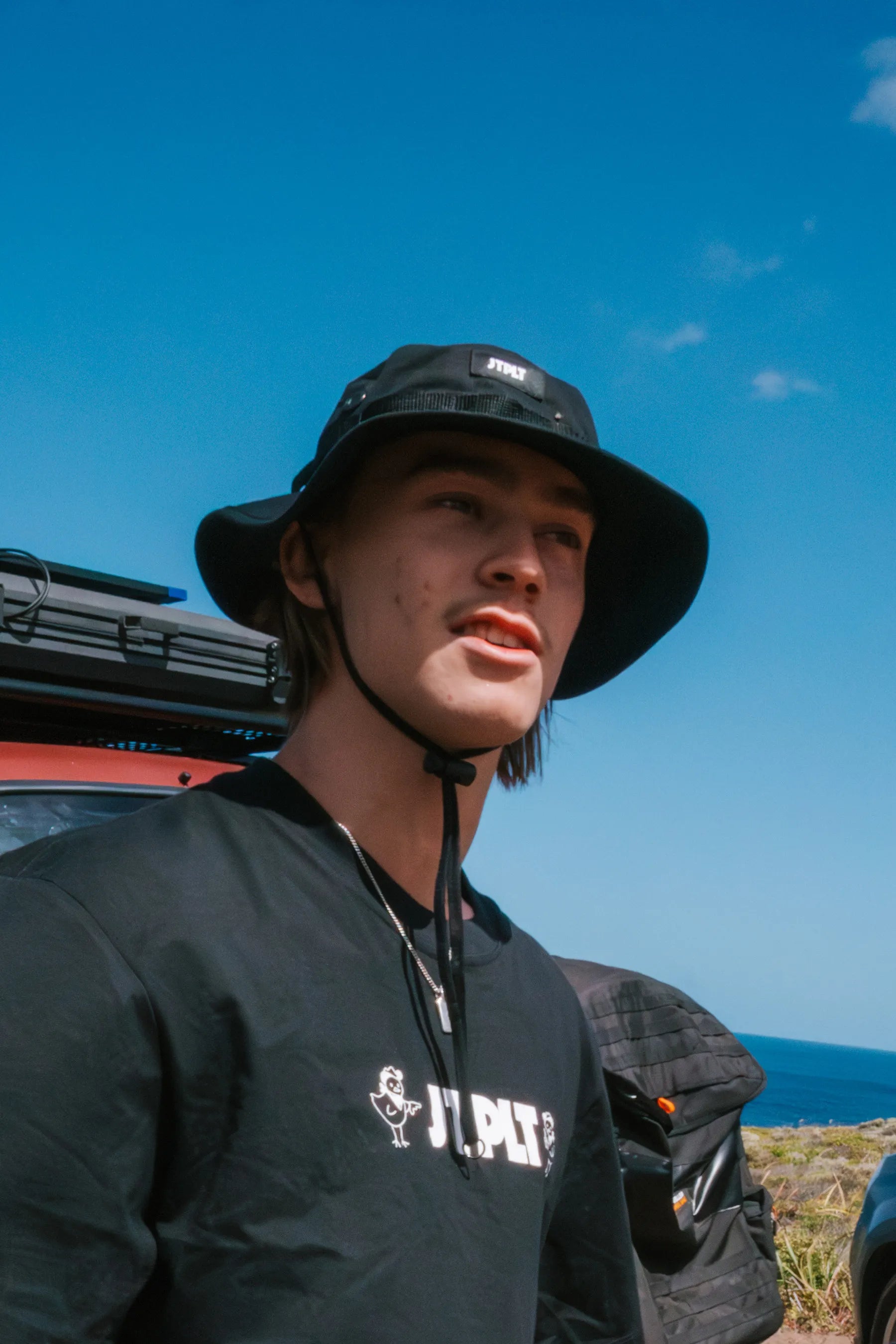 Person wearing a black bucket hat and dark t-shirt with visible branding, standing outdoors against a clear blue sky and beac