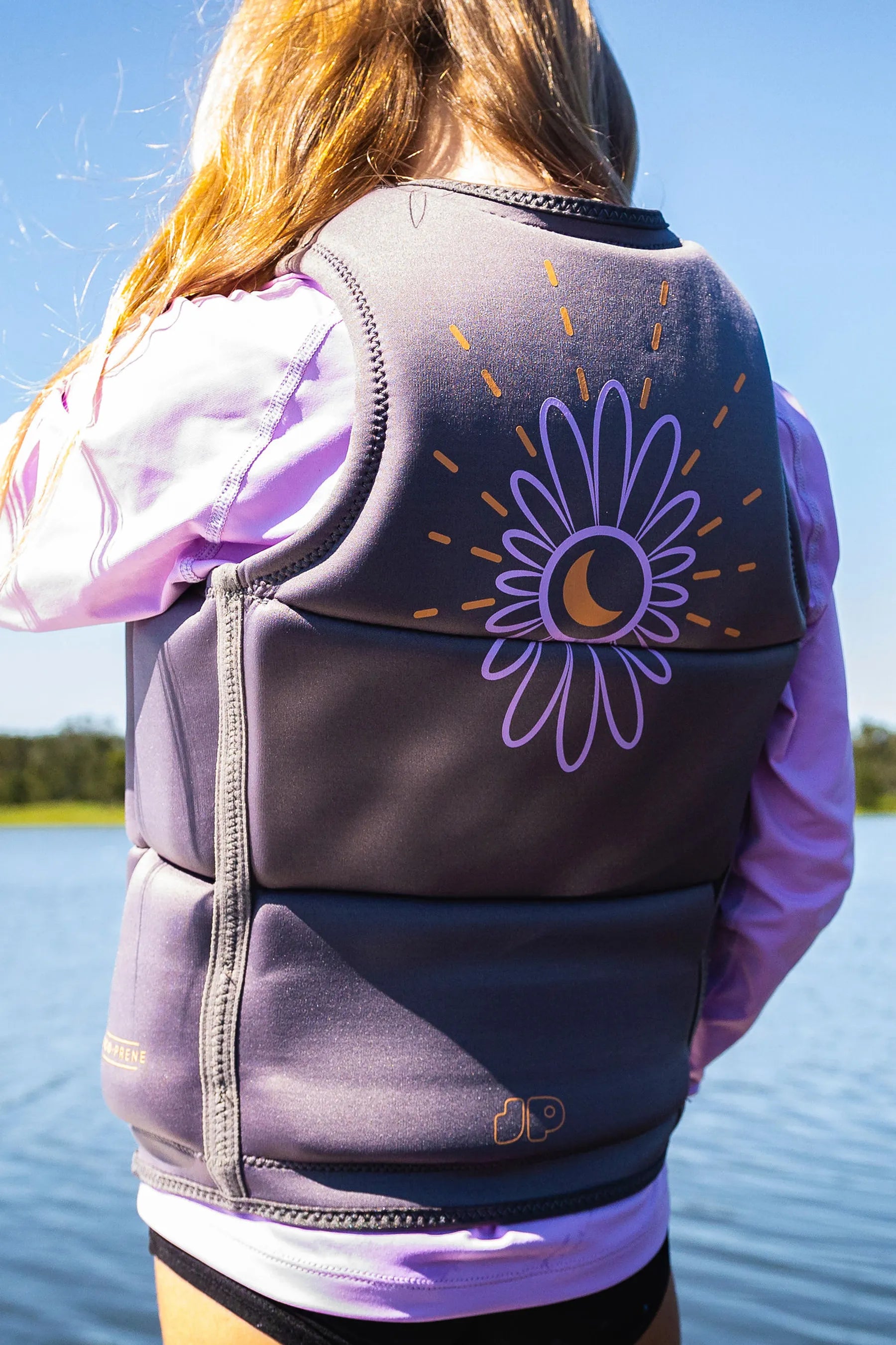 Girl in Jetpilot Freeride Girls Life Jacket - Charcoal, back view of vest with floral graphic, with purple long sleeve rash shirt underneath, on a pontoon by a lake