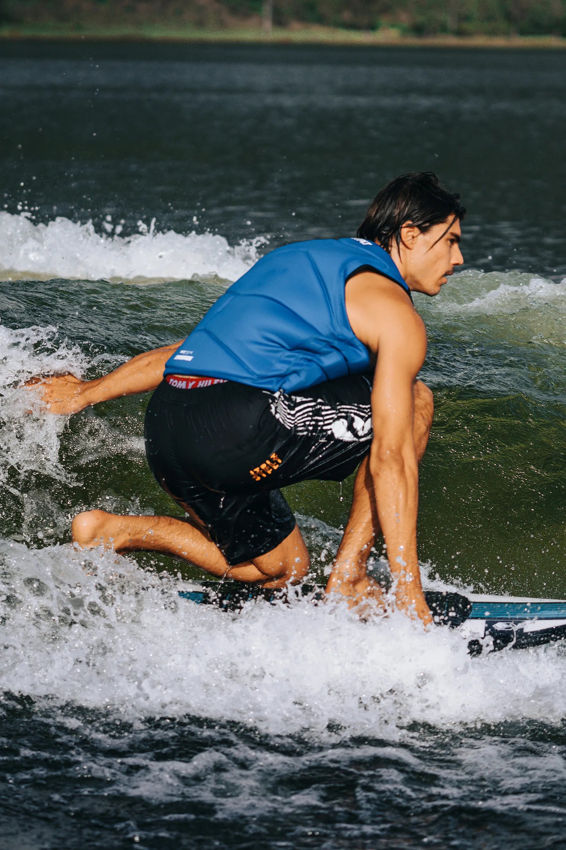Person wake surfing on a lake with a blue life jacket and black shorts.