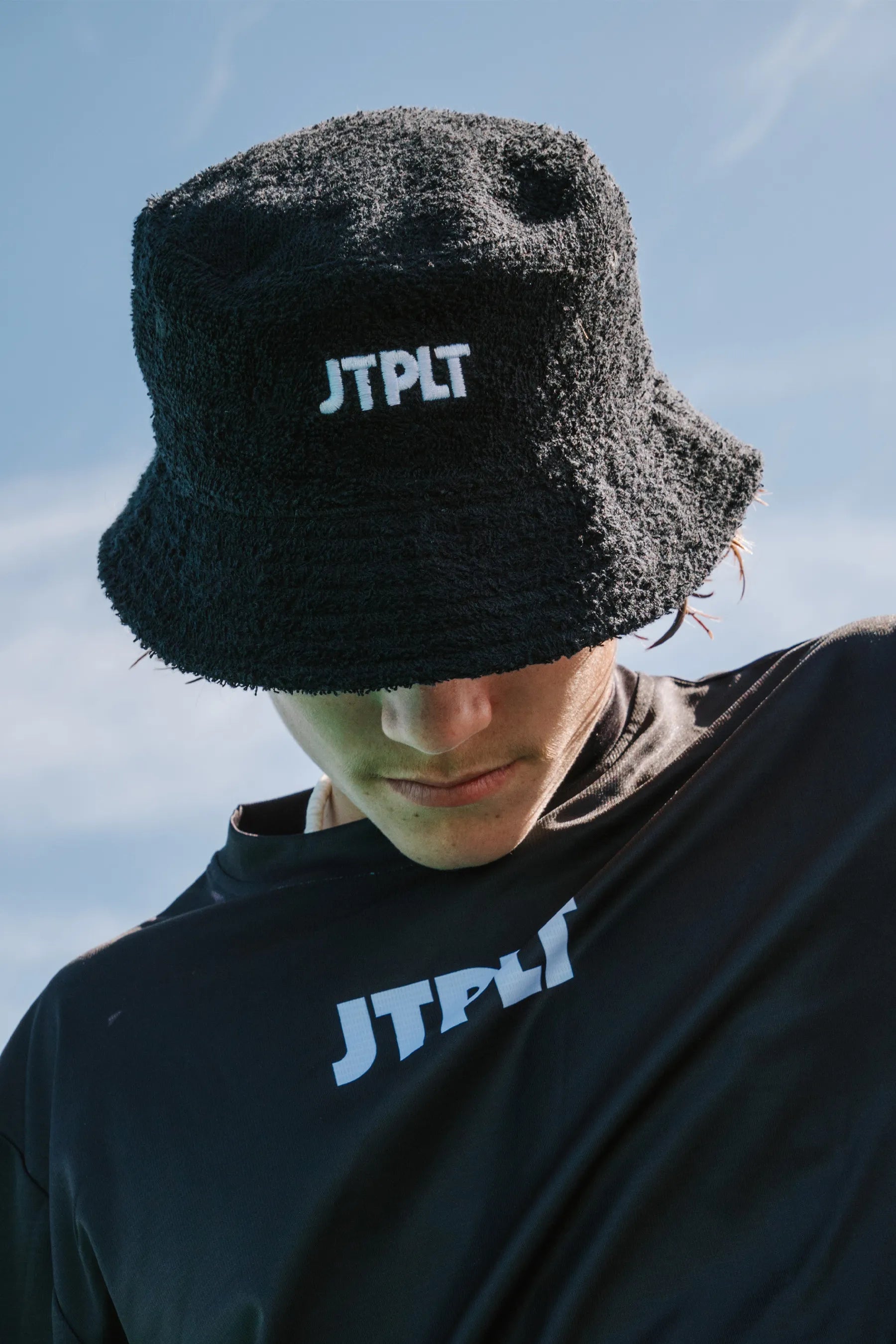 Person wearing a black bucket hat and black shirt with 'JTPLT' logo against a blue sky.