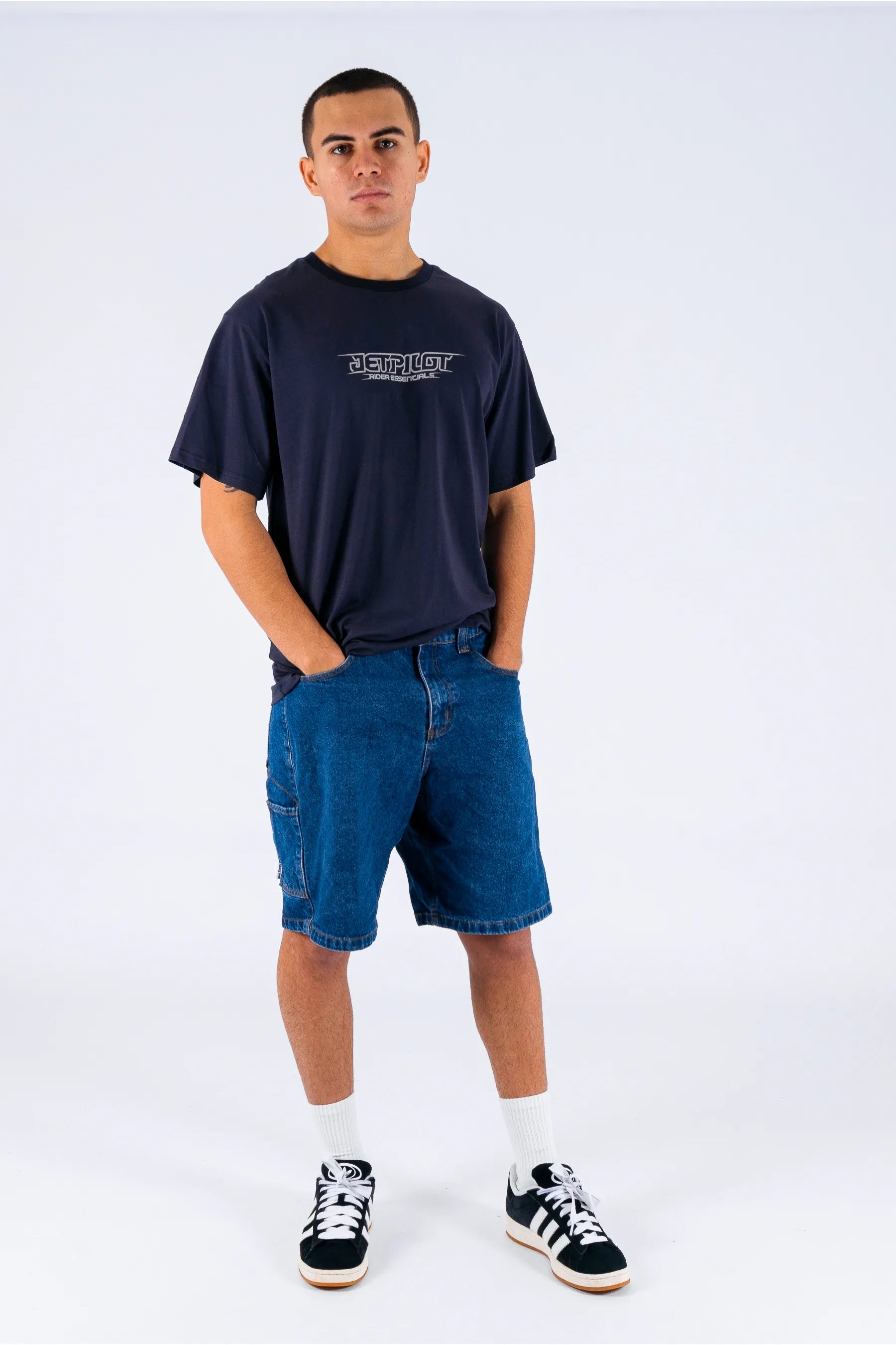 a person wearing a dark blue tee with a Jetpilot logo, blue shorts and a black/white shoes on a white background — view 5 (3)