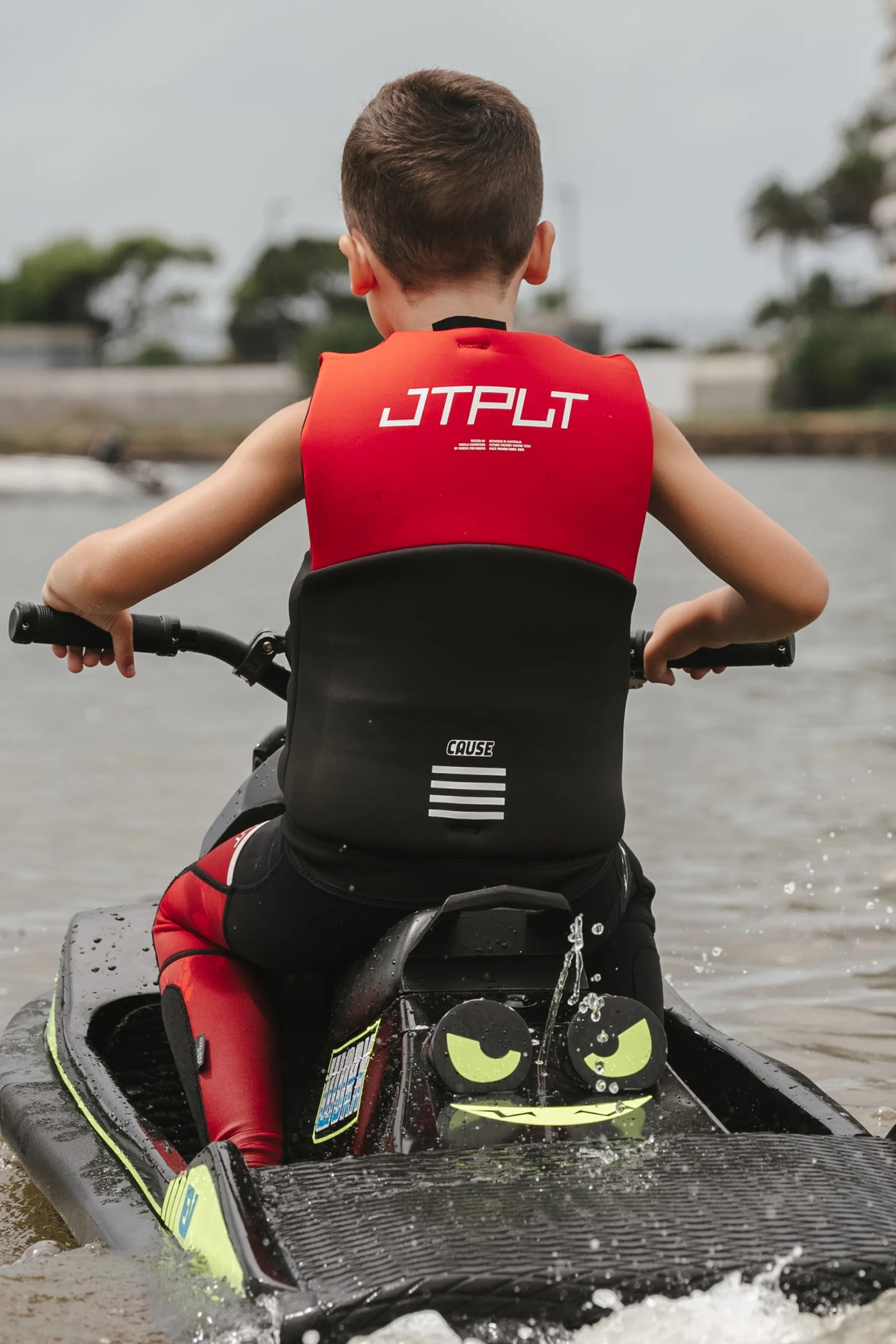 Child wearing a Cause Kids Life Jacket - Red L50 on a watercraft with a scenic background.