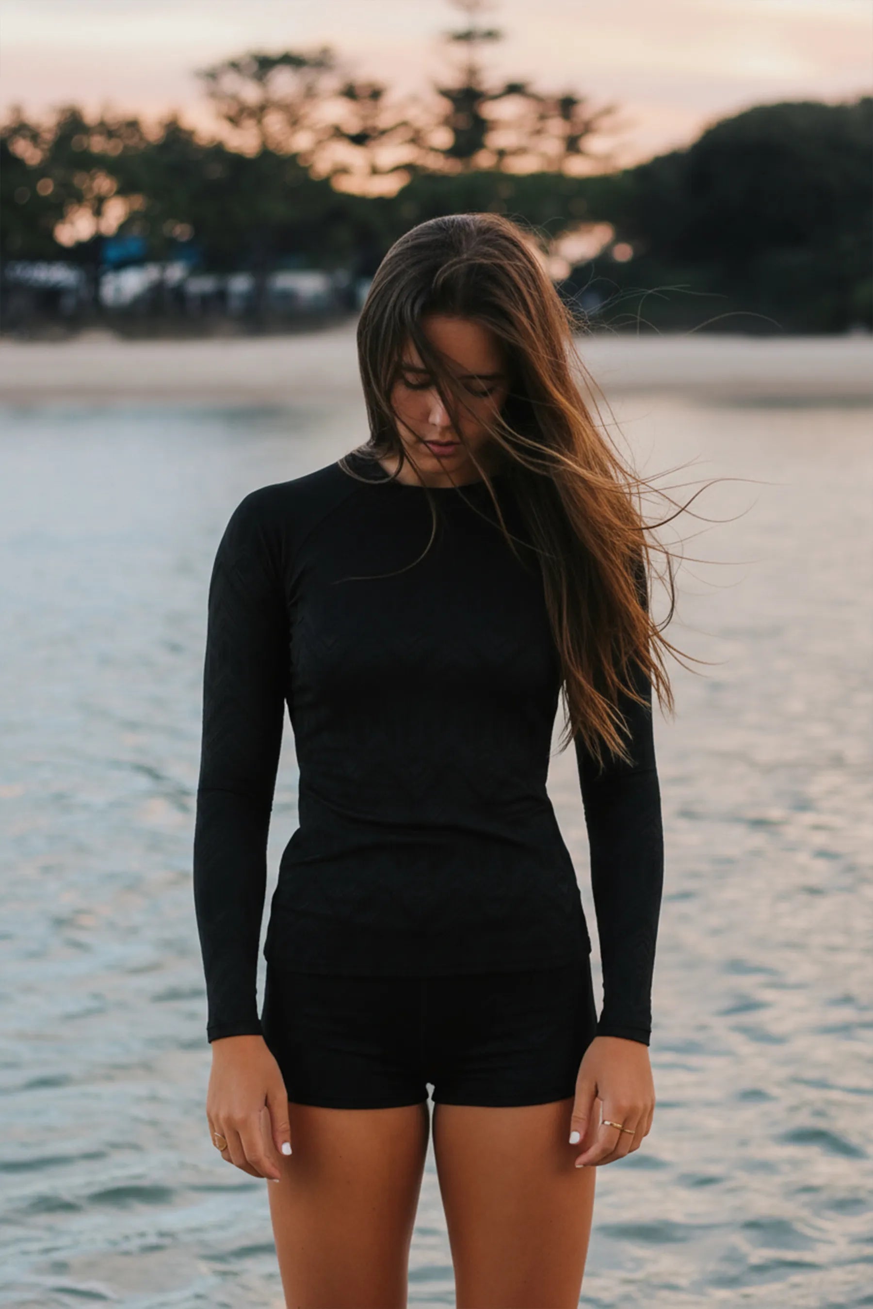 Woman wearing the Jetpilot Rash Shirt & Short Set - Black standing by a body of water with trees in the background. — view 1