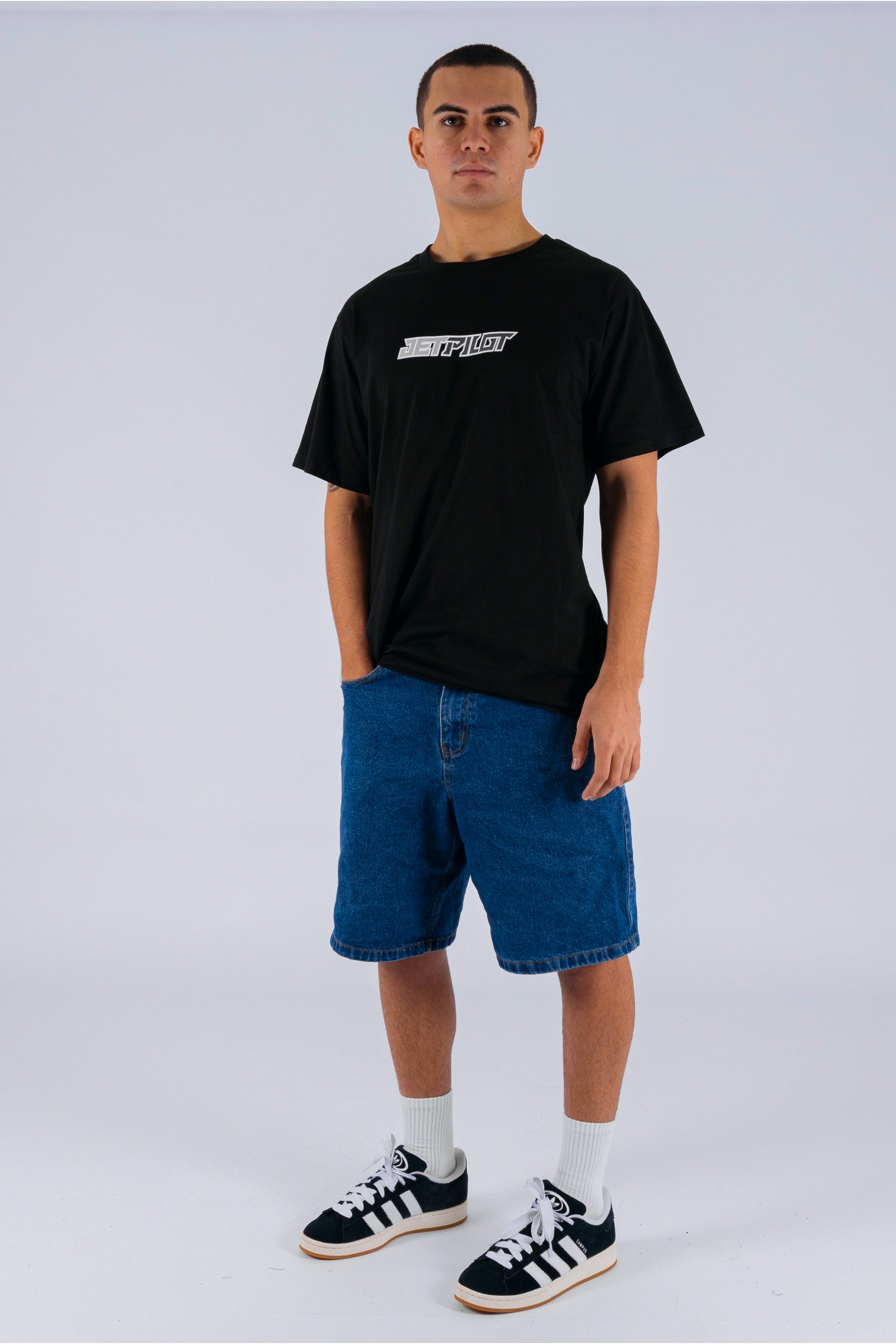 A person wearing a black tee with a Jetpilot logo, blue denim shorts and black/white shoes. — view 3 (3)
