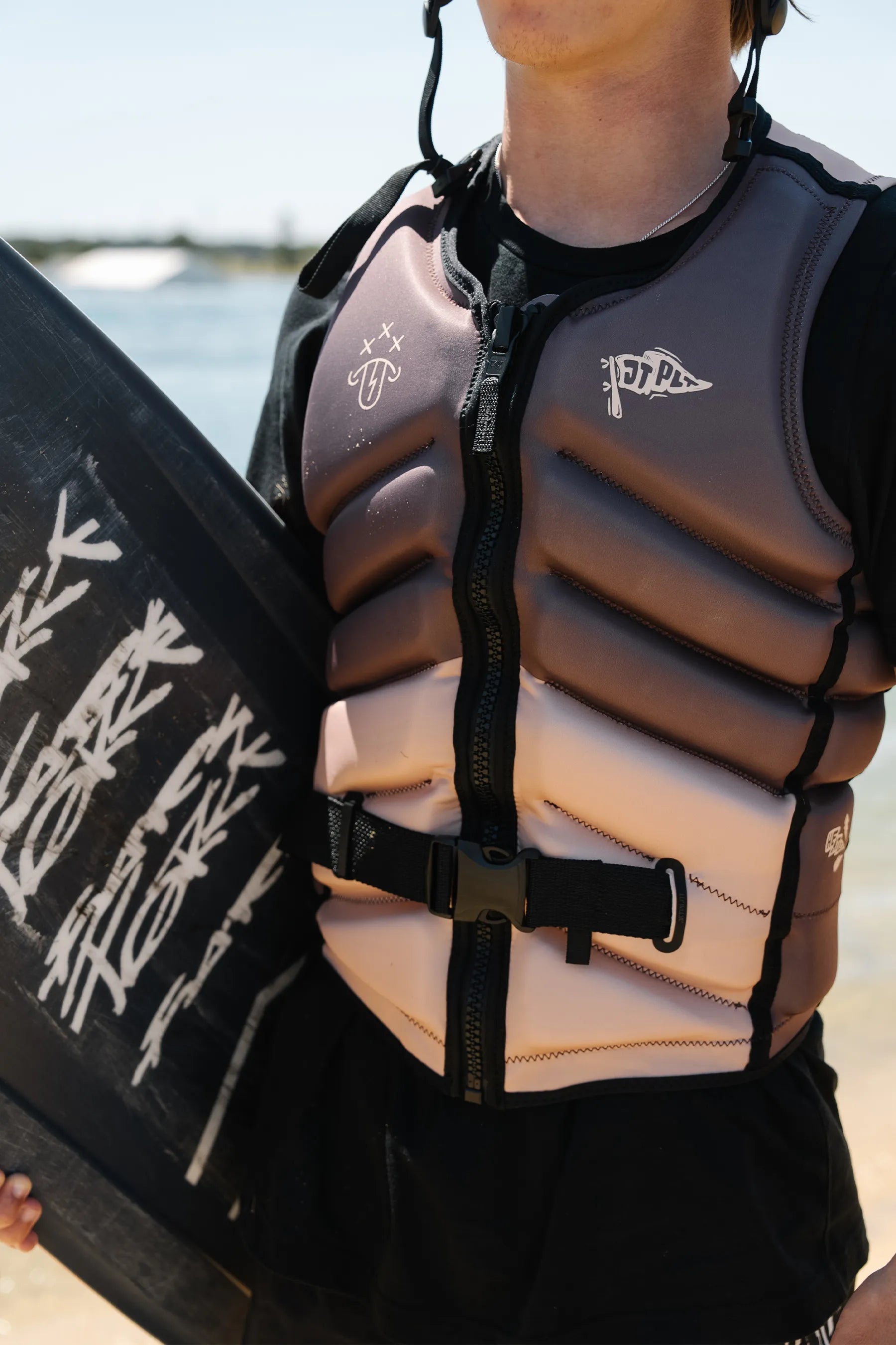 Person wearing a Jetpilot X1 Life Jacket - JTPLT X Heatley - Brown holding a wakeboard with text on a beach.