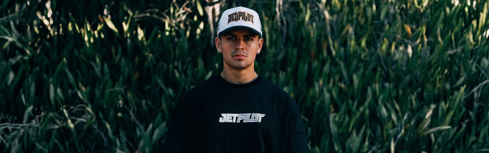 A man wearing a white Jetpilot snapback and an all black t-shirt with a Grey and White "Jetpilot" logo against an outdoor garden
