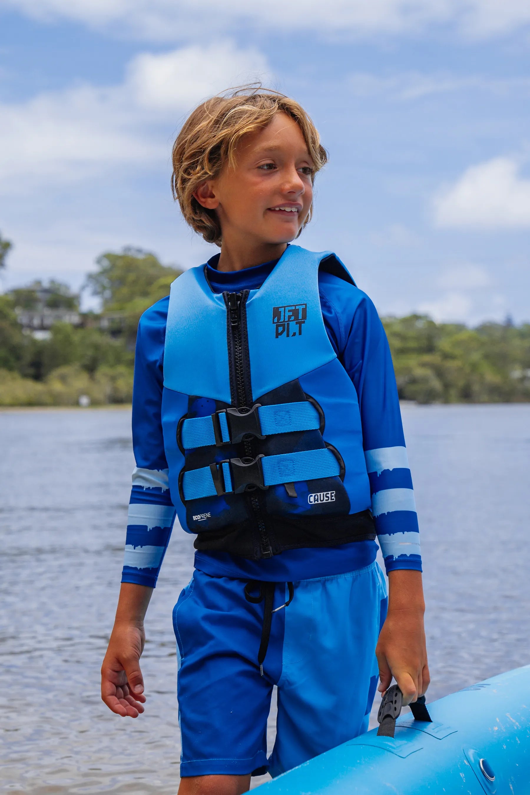 Child wearing a blue life jacket and shorts by a body of water.