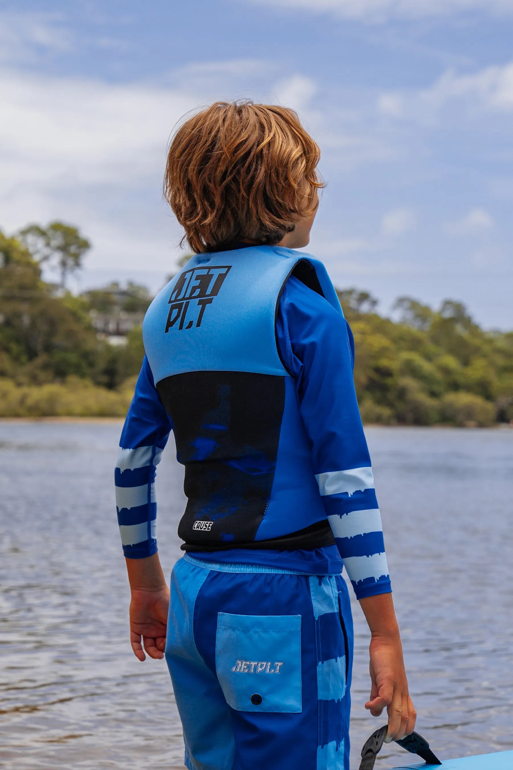 Child wearing a blue and black life jacket by a body of water with trees in the background