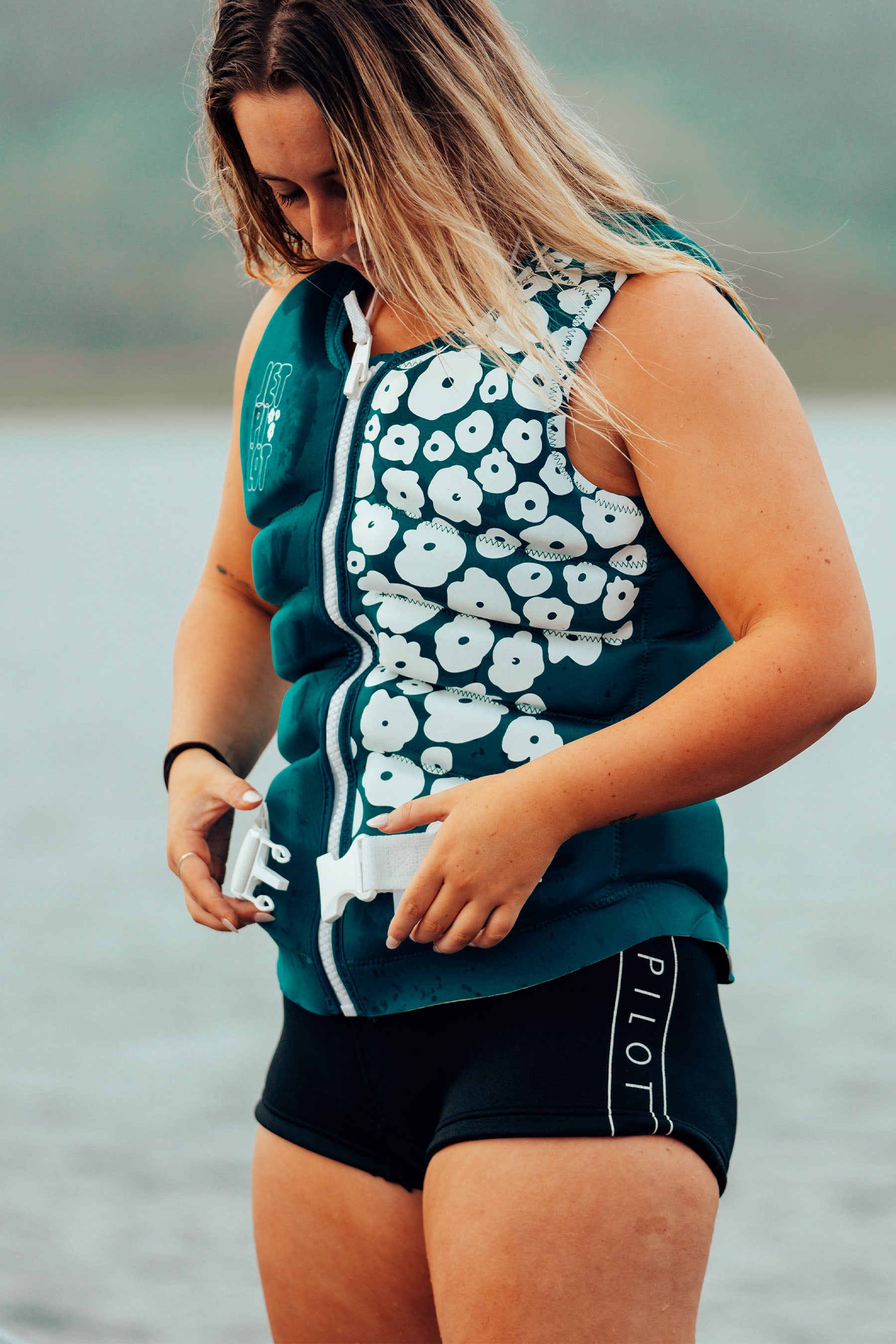 Person wearing a floral-patterned life jacket and black shorts by a body of water.