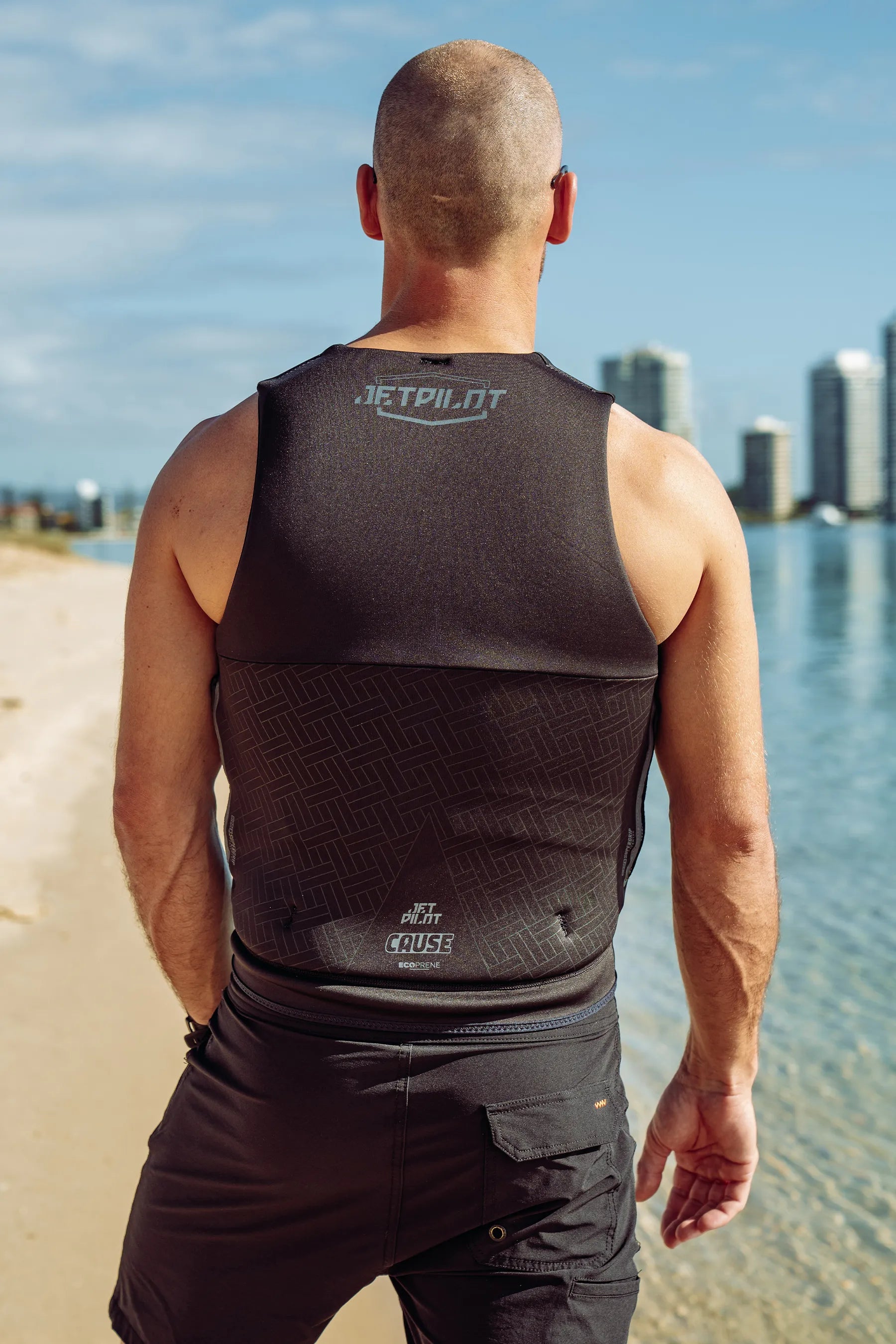 Man wearing a Jetpilot Cause Life Jacket - Black with 'JETPILOT' branding on a beach.