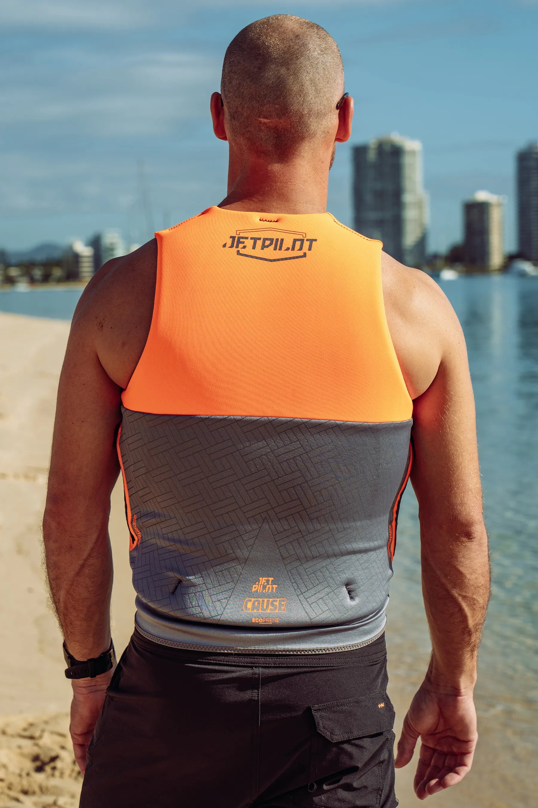 Man wearing a Jetpilot Cause Life Jacket - Charcoal/Orange L50 on a beach with buildings in the background