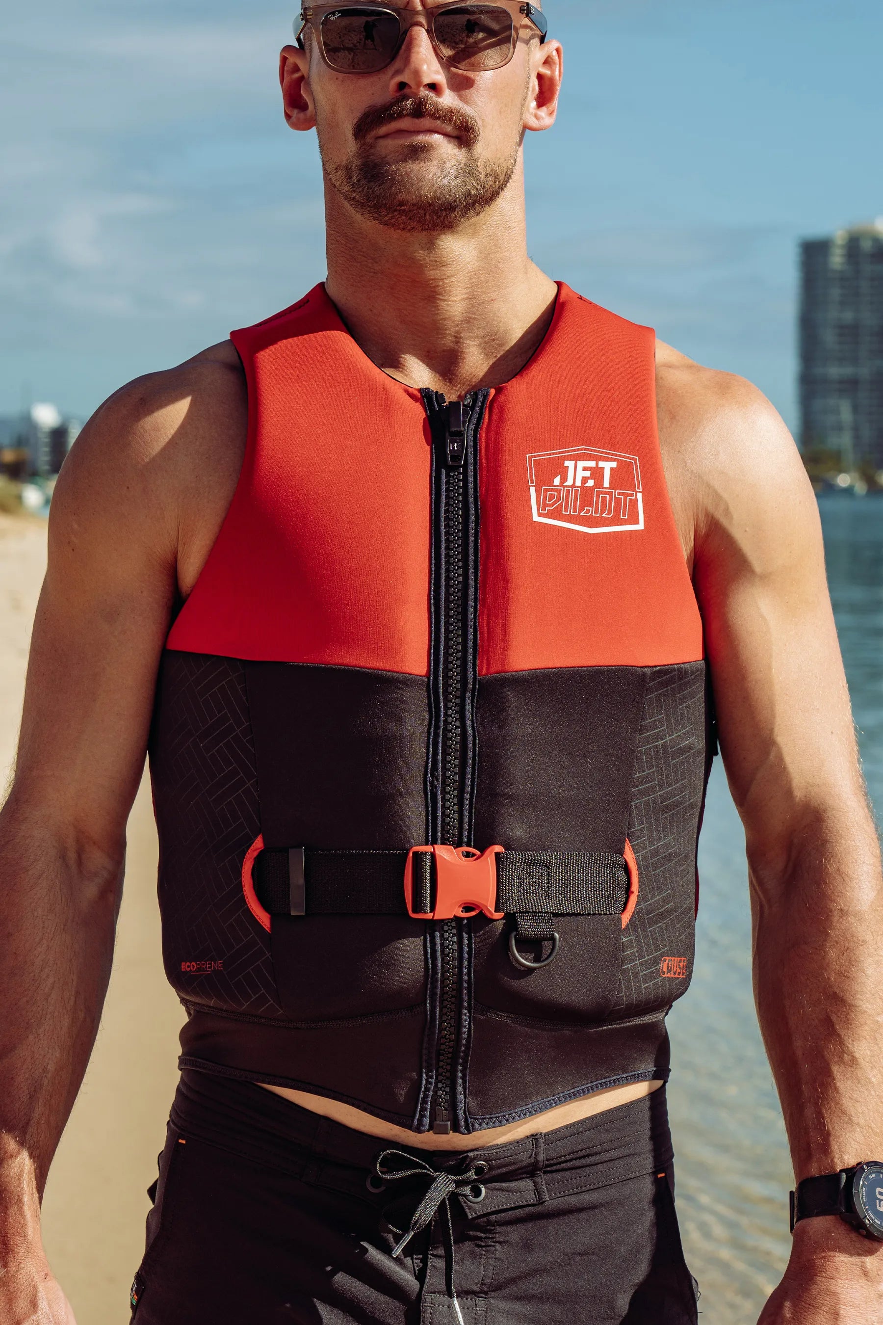 Man wearing a Jetpilot Cause Life Jacket - Red L50 with a logo on a beach.