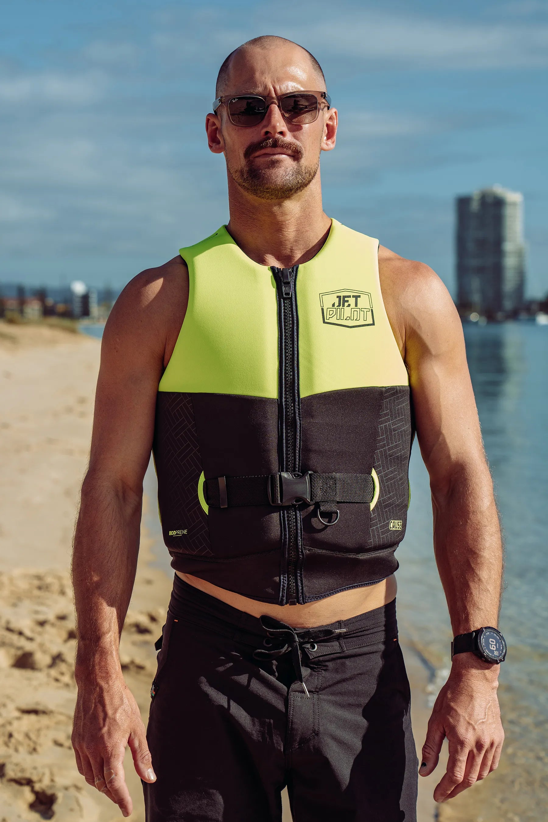 Man wearing a Jetpilot Cause Life Jacket - Yellow L50 on a beach with water and buildings in the background