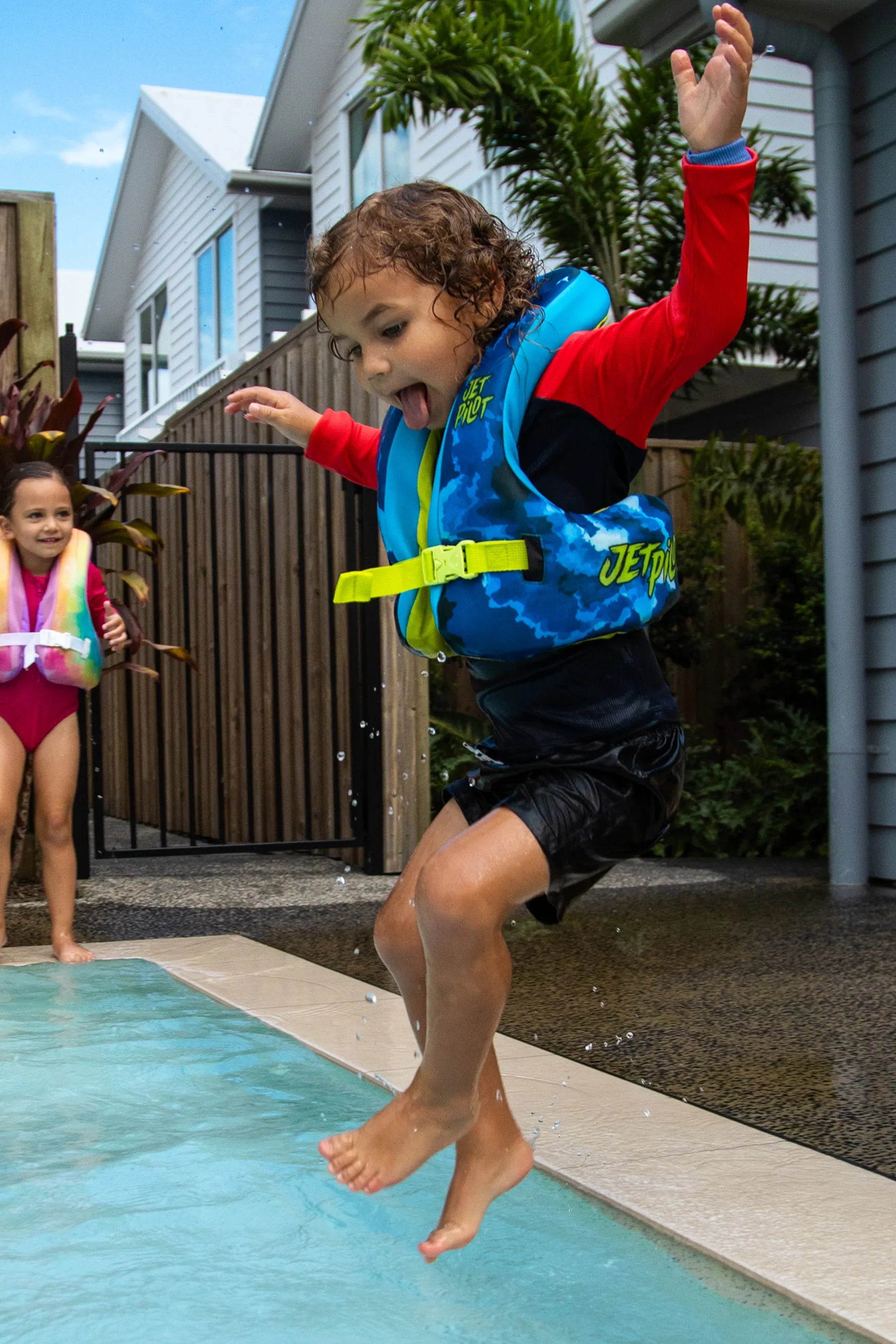 Child in a life jacket jumping into a pool with another child watching.