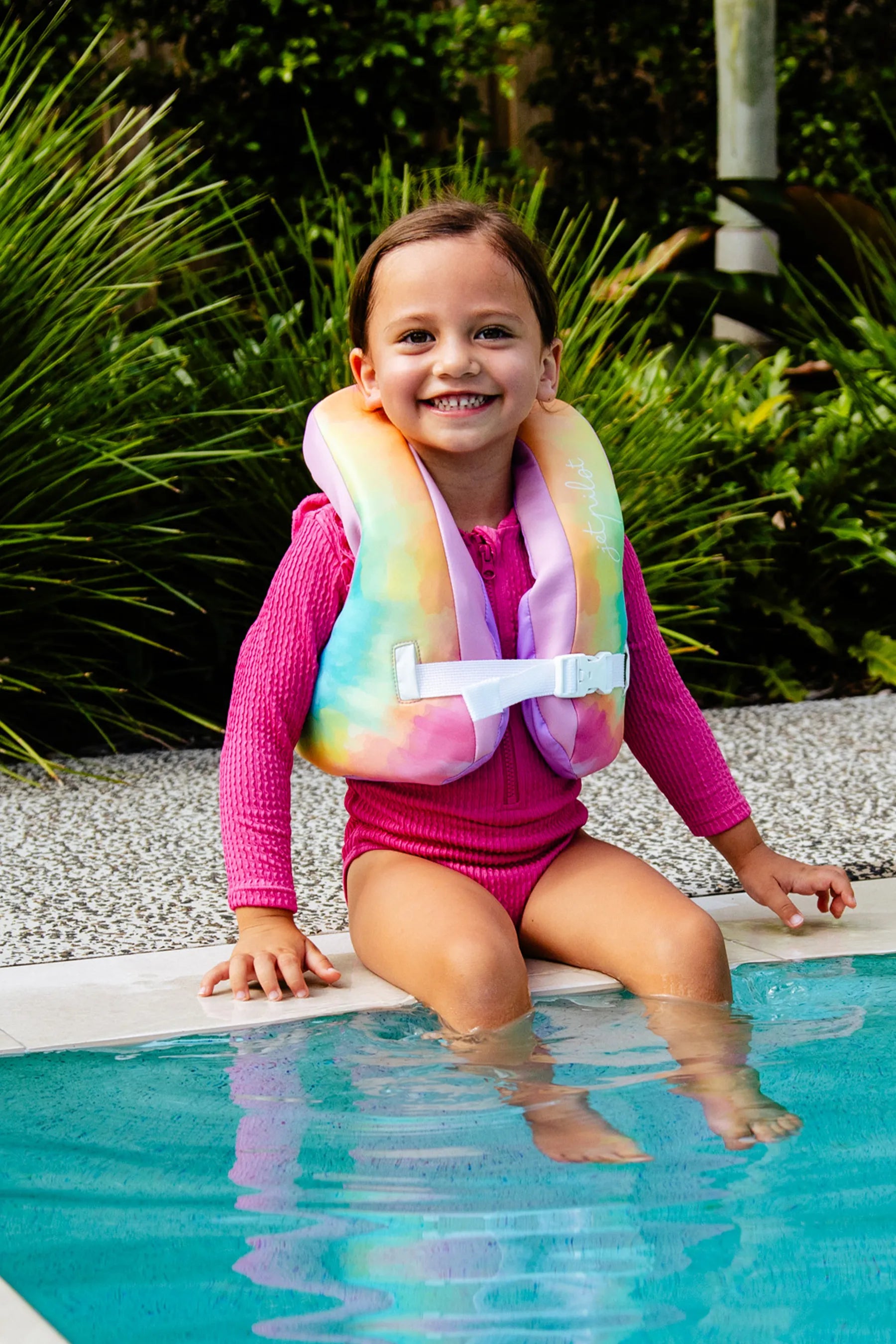 Child wearing a colorfulJ etpilot Paddle Pal Splash Swim Vest - Tie Dye sitting by a pool with greenery in the background