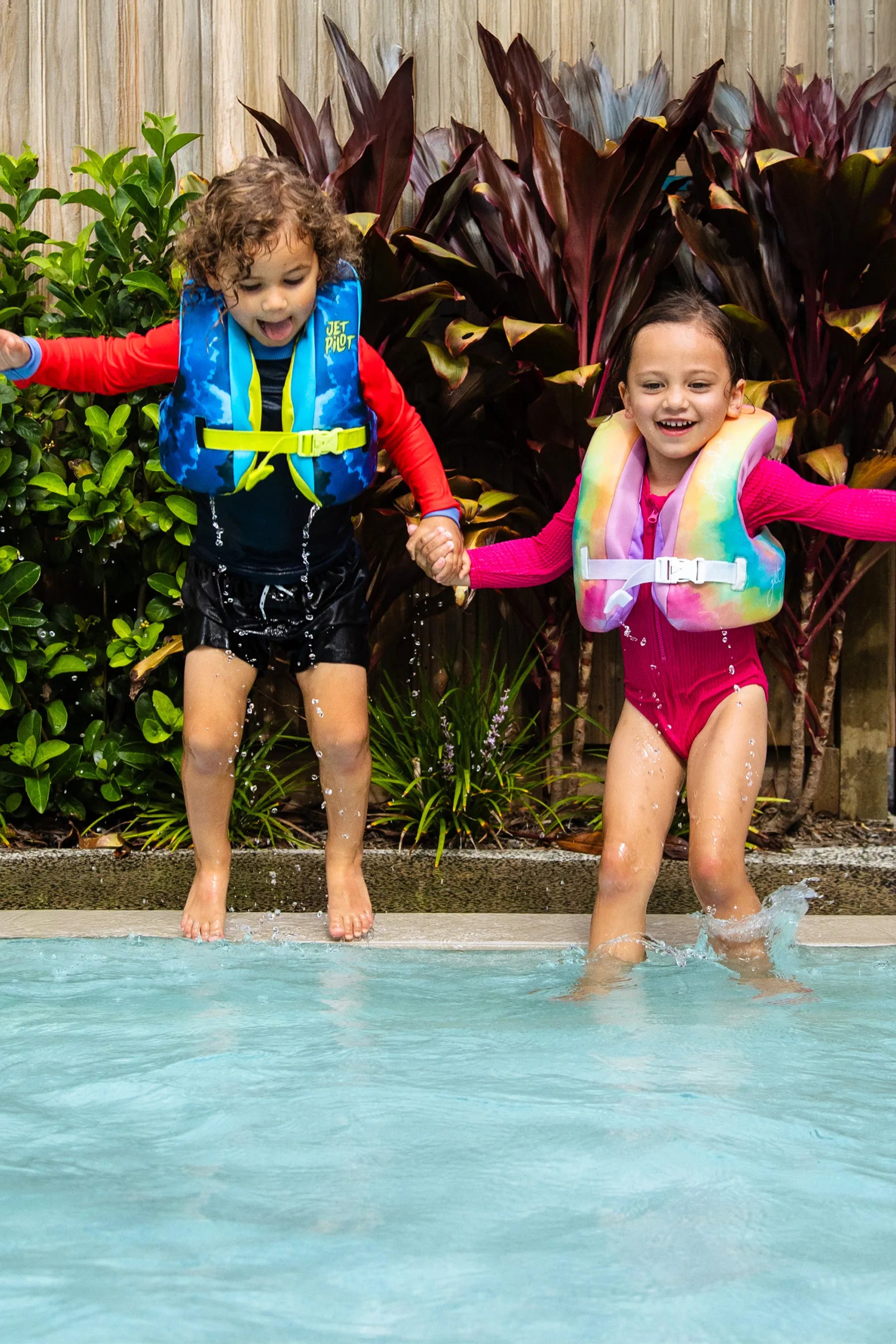 Two children in Jetpilot Paddle Pals life jackets holding hands as they jump into a pool.