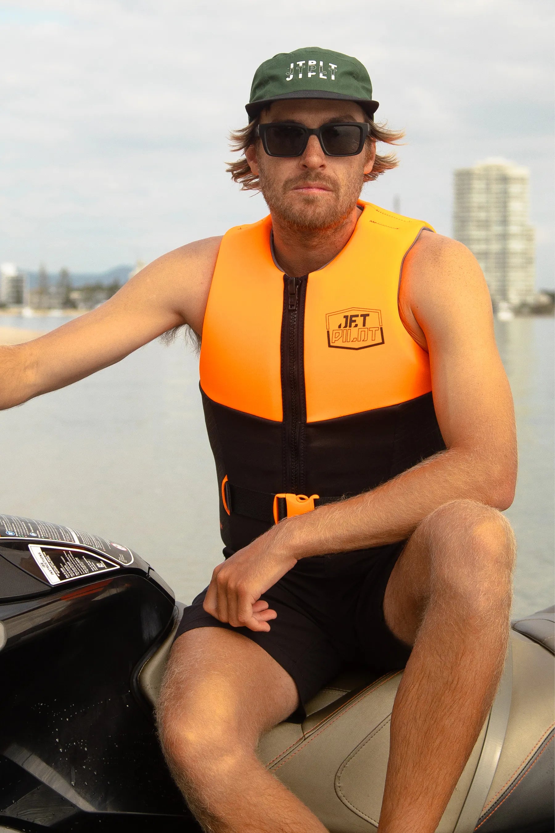 Man wearing an orange life vest and sunglasses sitting on a boat with a cityscape in the background.