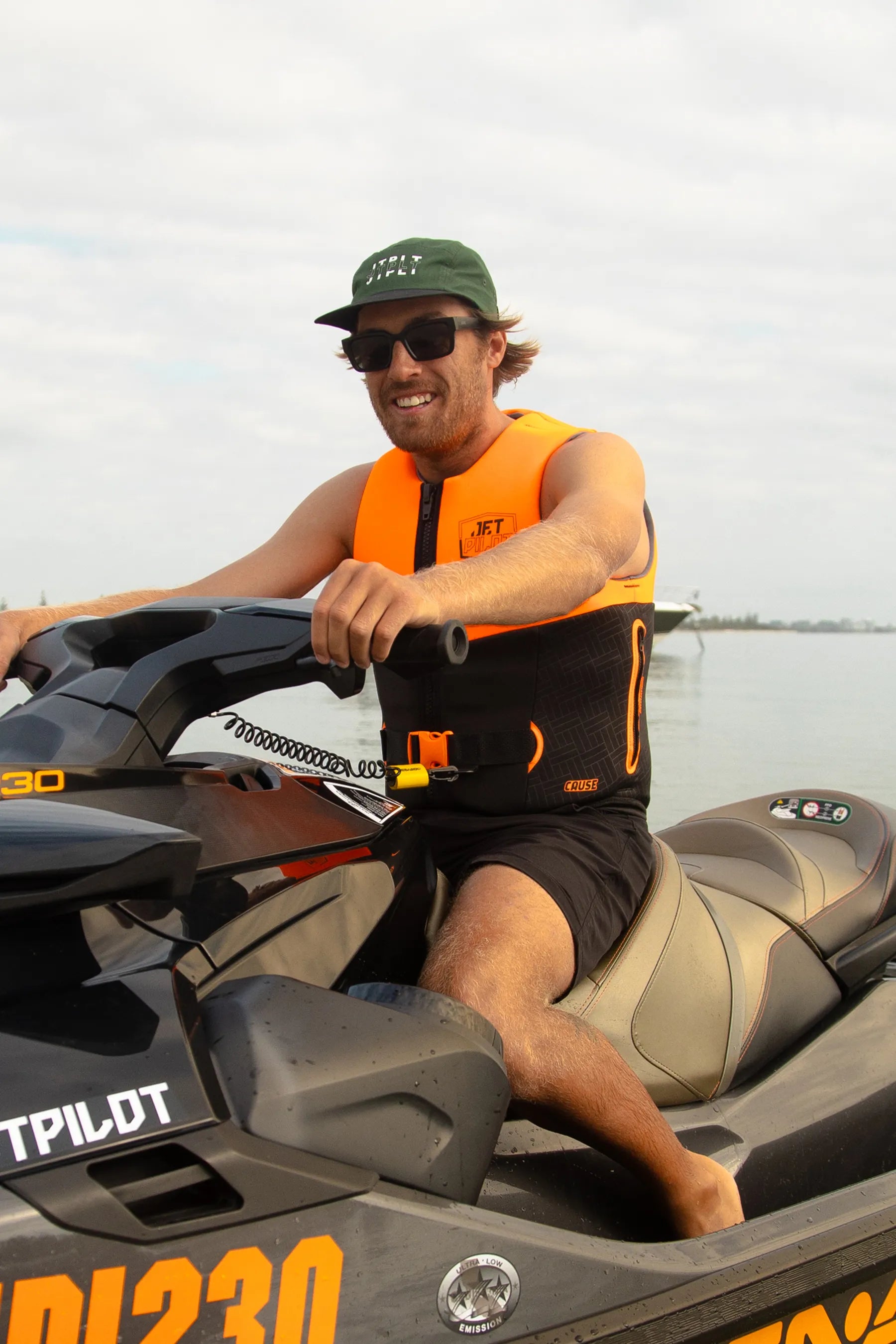 Man on a jet ski wearing an orange life vest and sunglasses, with water and sky in the background.