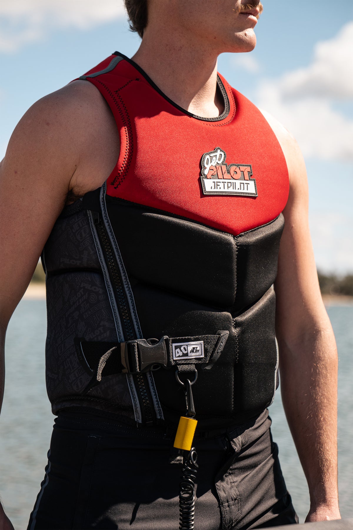 A close-up of a man wearing a red and black Jetpilot life vest with a chest logo set against an outdoor water background.