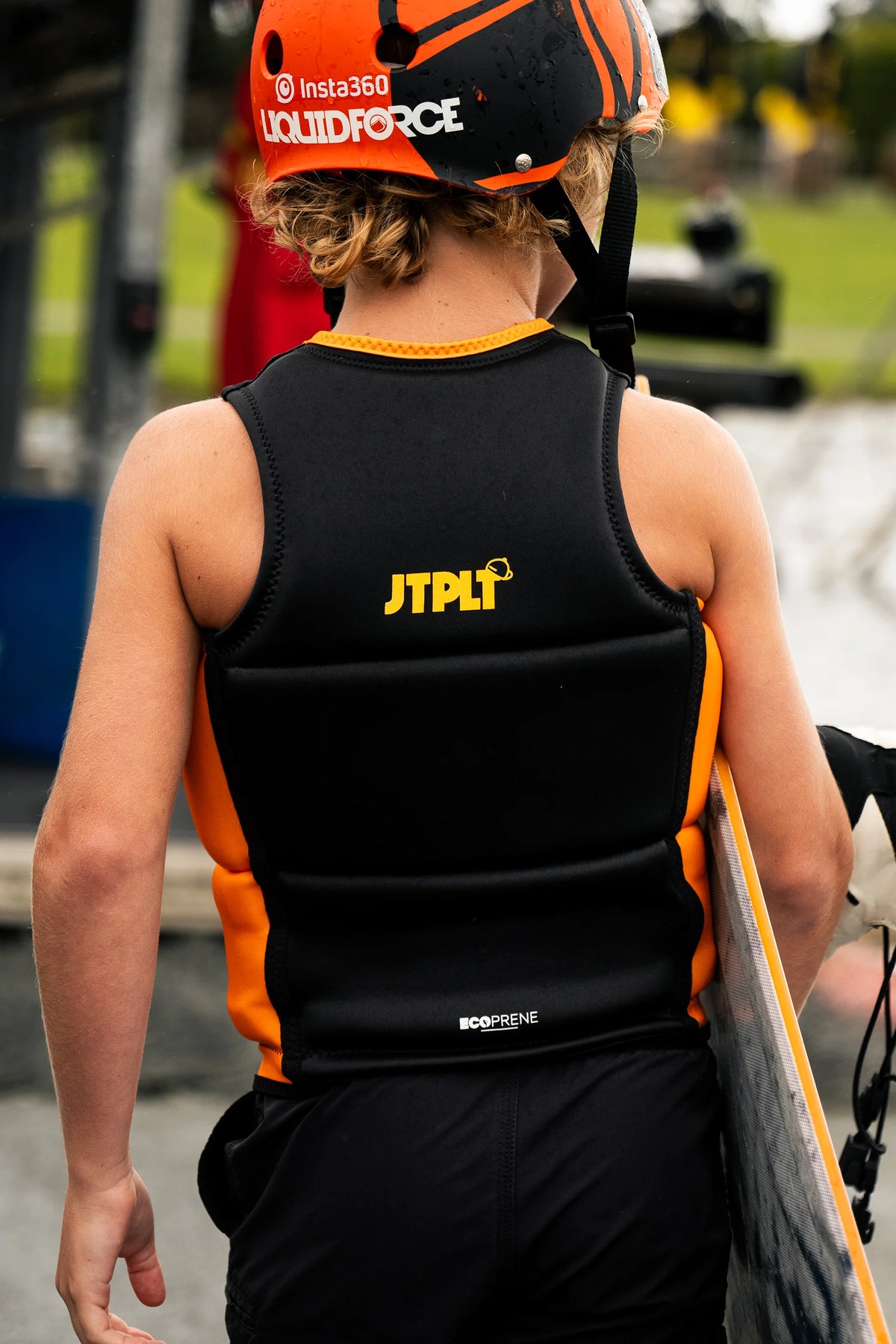 Young rider in a helmet walking away with a wakeboard, showing the JTPLT logo on the back of the Black Freeride Boys