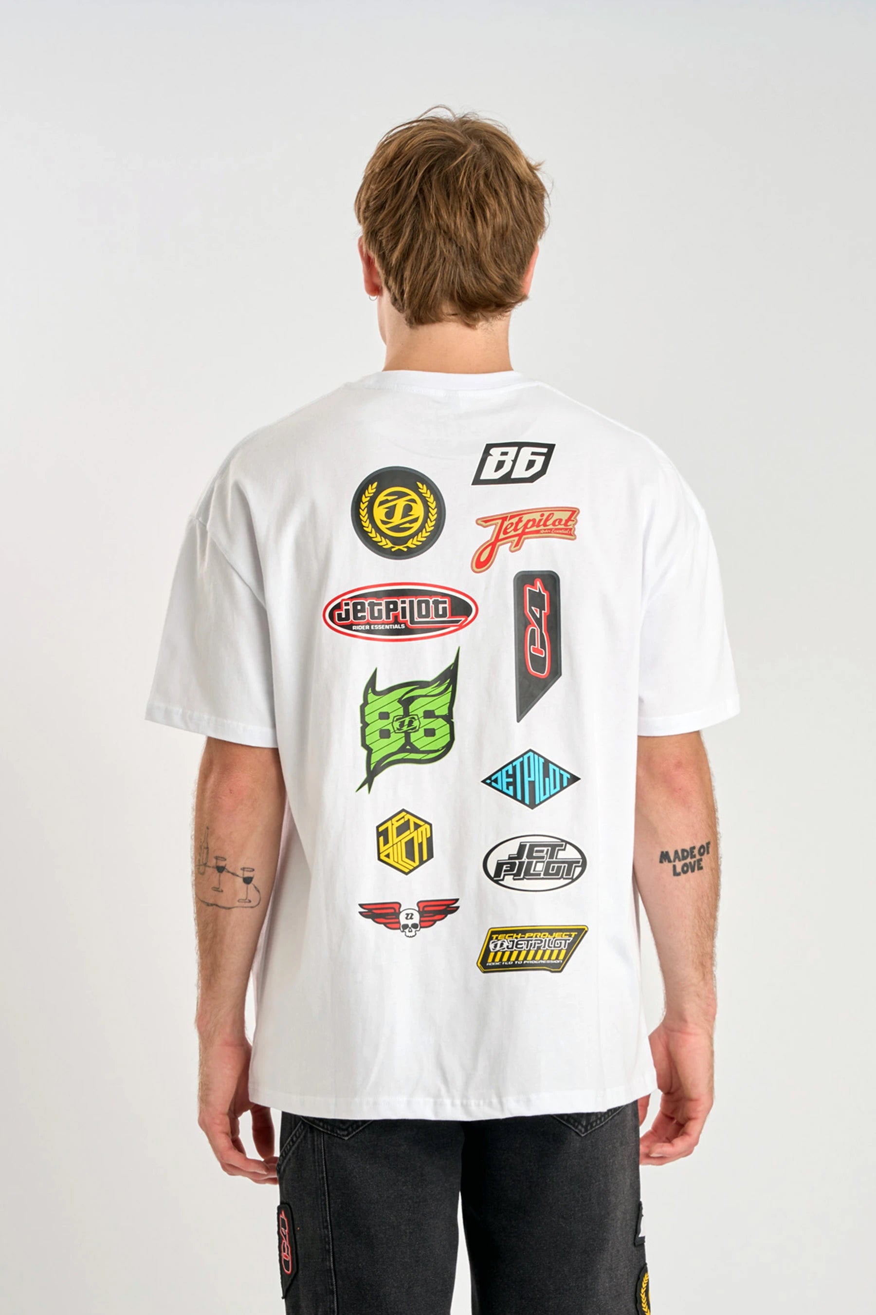 Person wearing a Jetpilot Factory Heavy Tee - White with various colorful logos on the back against a plain background — imag