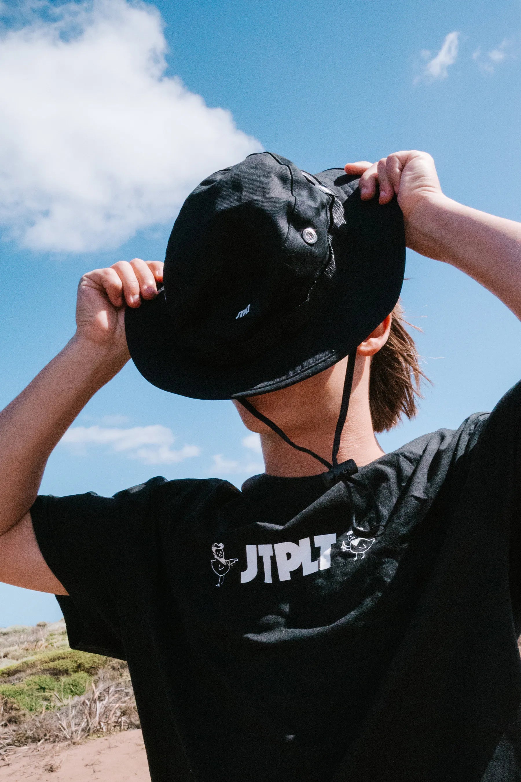 Person wearing a black bucket hat and black Jetpilot T-shirt with visible branding against a blue sky. — image 2