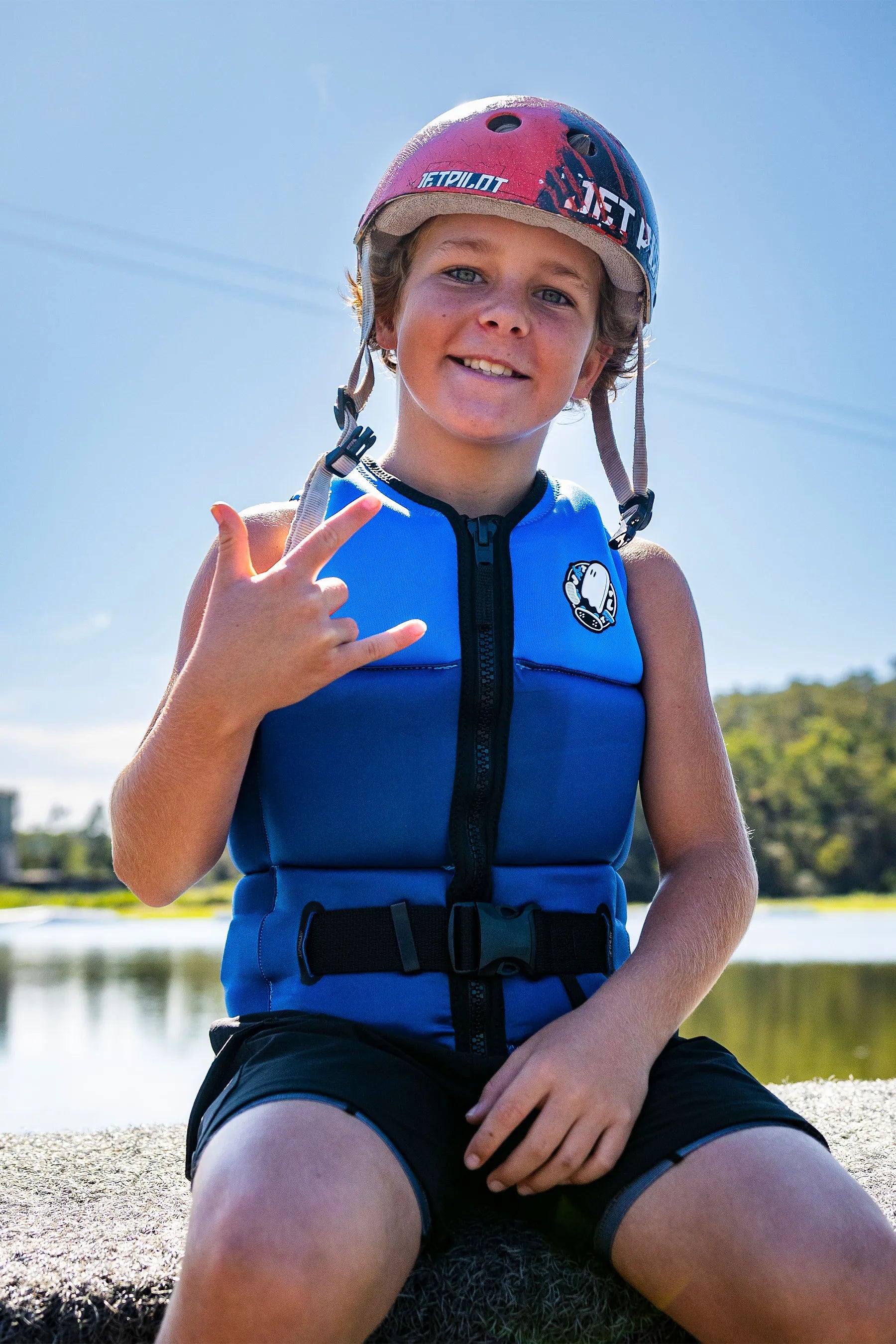 Child wearing a Jetpilot Freeride Boys Life Jacket - Blue and pink helmet by a lake
