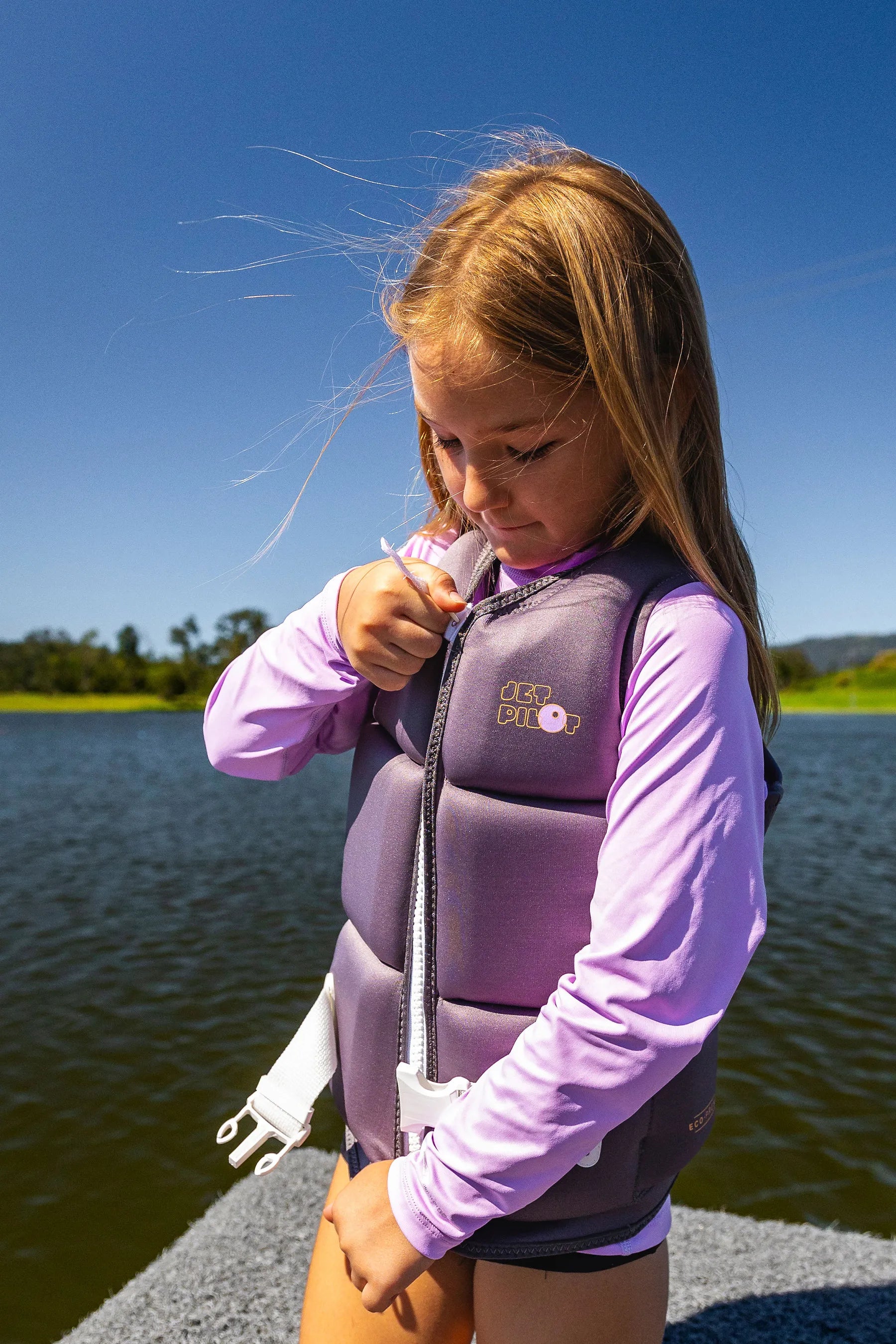 Girl in Jetpilot Freeride Girls Life Jacket - Charcoal, with purple long sleeve rash shirt underneath, on a pontoon by a lake