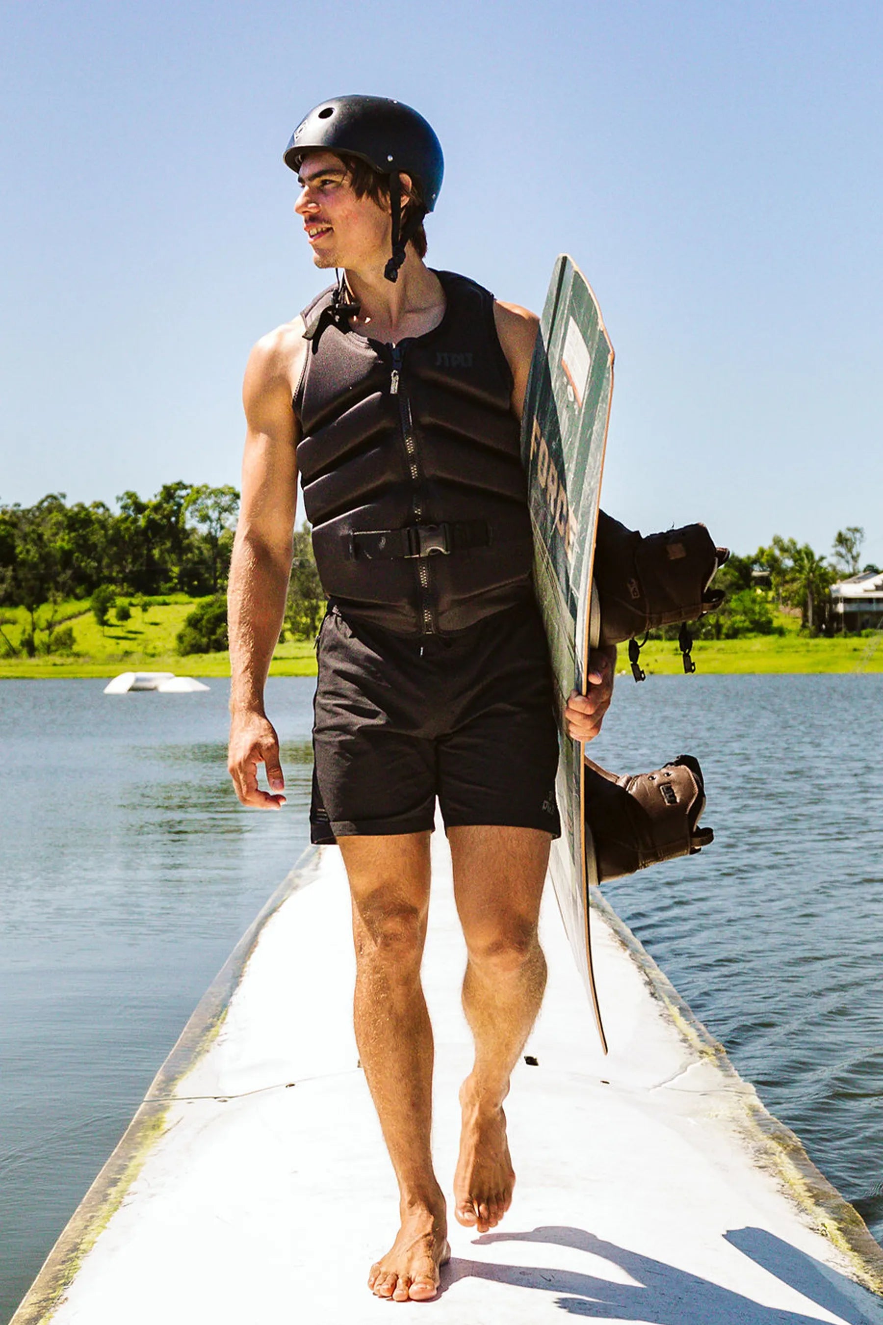Person in Jetpilot Freeride Life Jacket - Black holding a wakeboard on a dock by a lake.