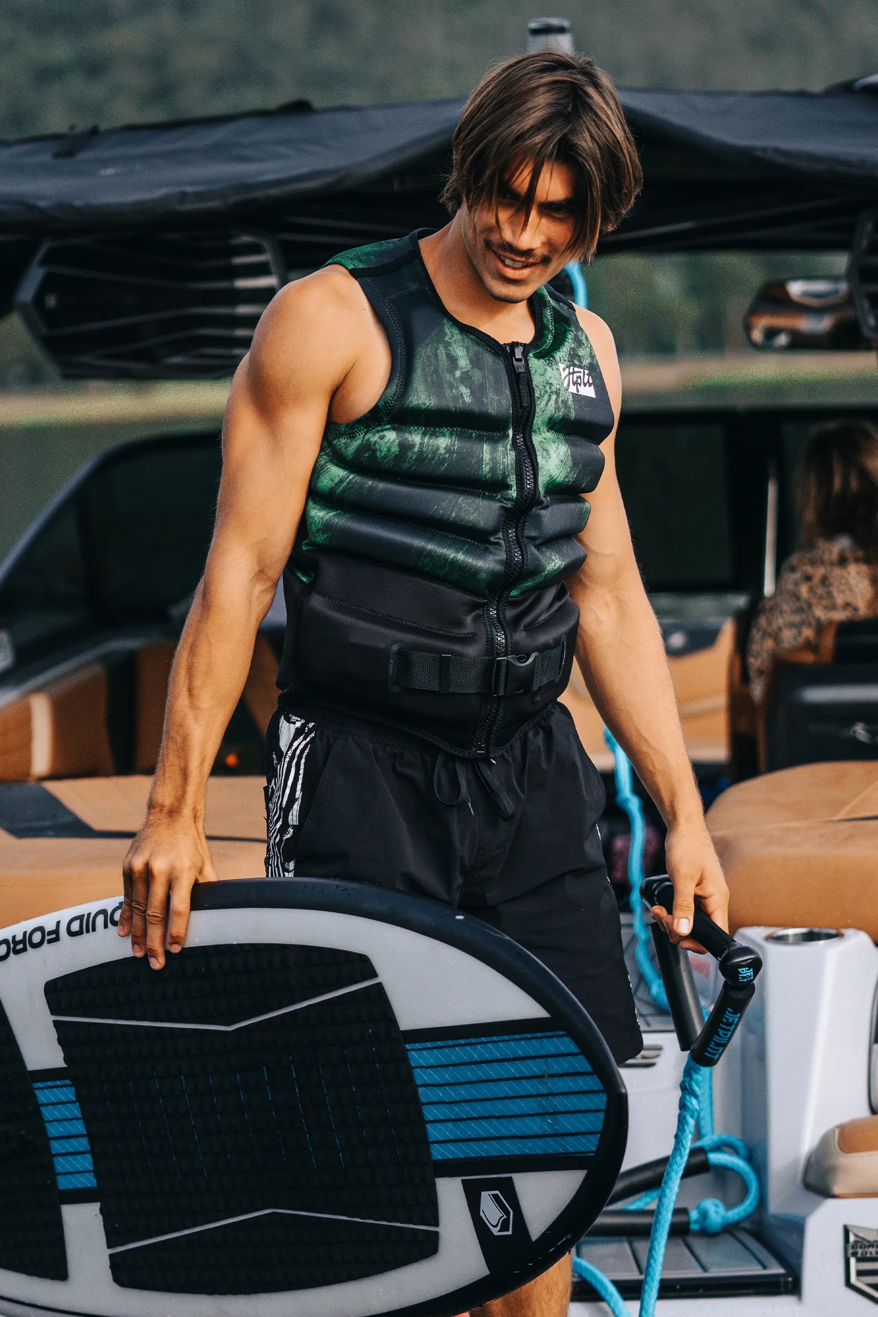Person wearing a Jetpilot Phase Hyperflex Life Jacket - Green holding a wakeboard on a boat