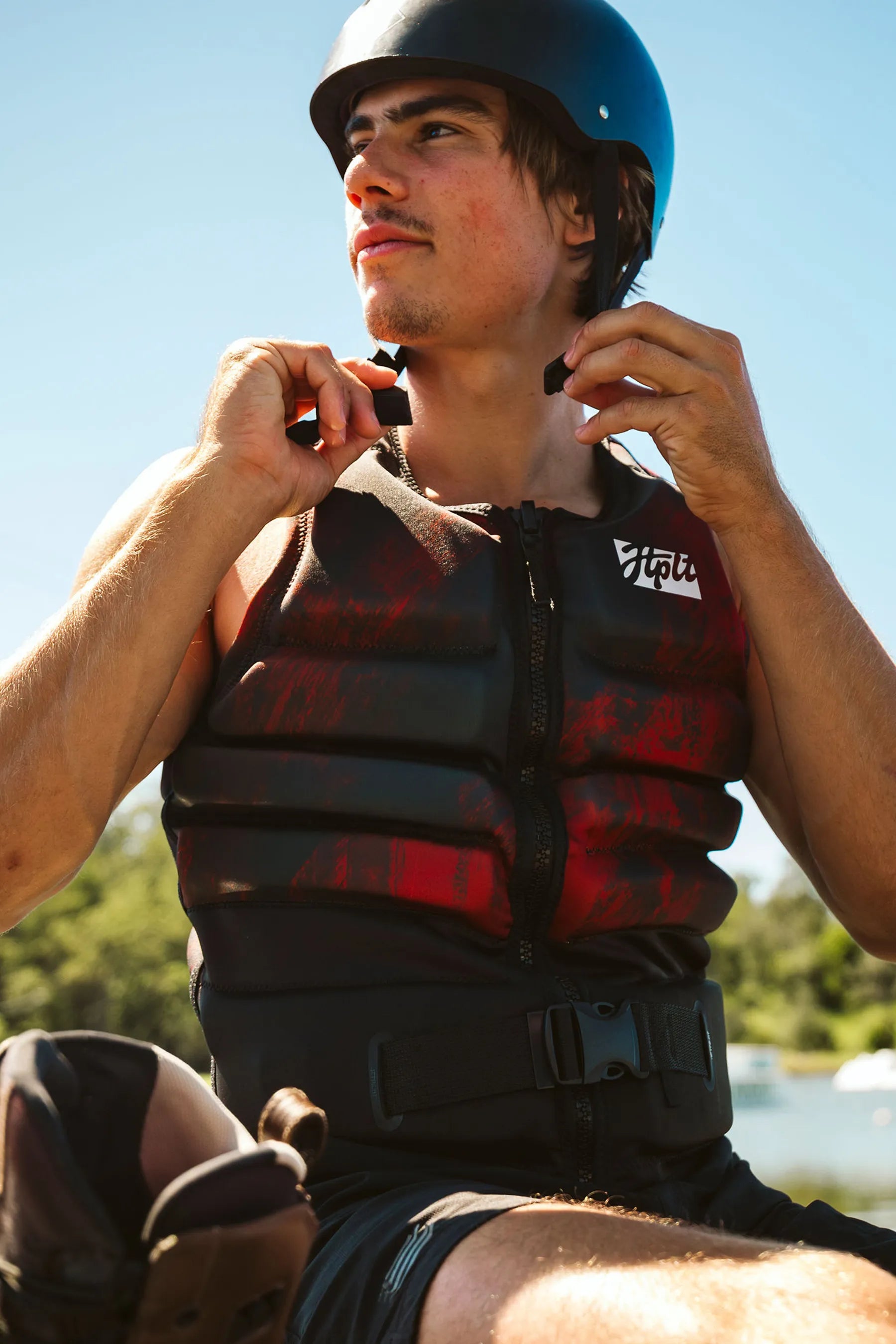 Person wearing a Jetpilot Phase Hyperflex Life Jacket - Red and helmet, sitting outdoors with a clear sky.