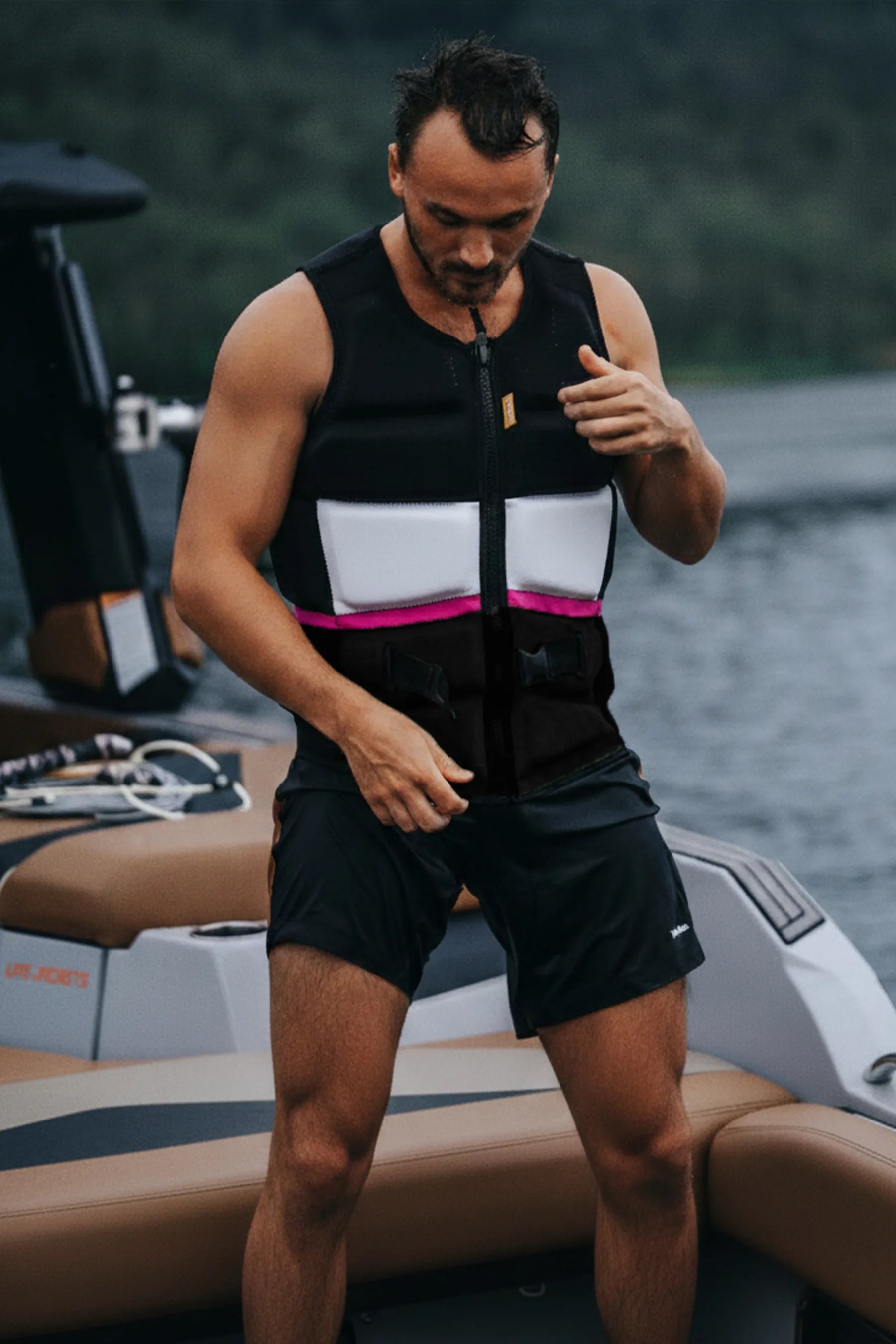 Man adjusting a Jetpilot Pro C4 Life Jacket - Black/Grey on a boat with water and trees in the background