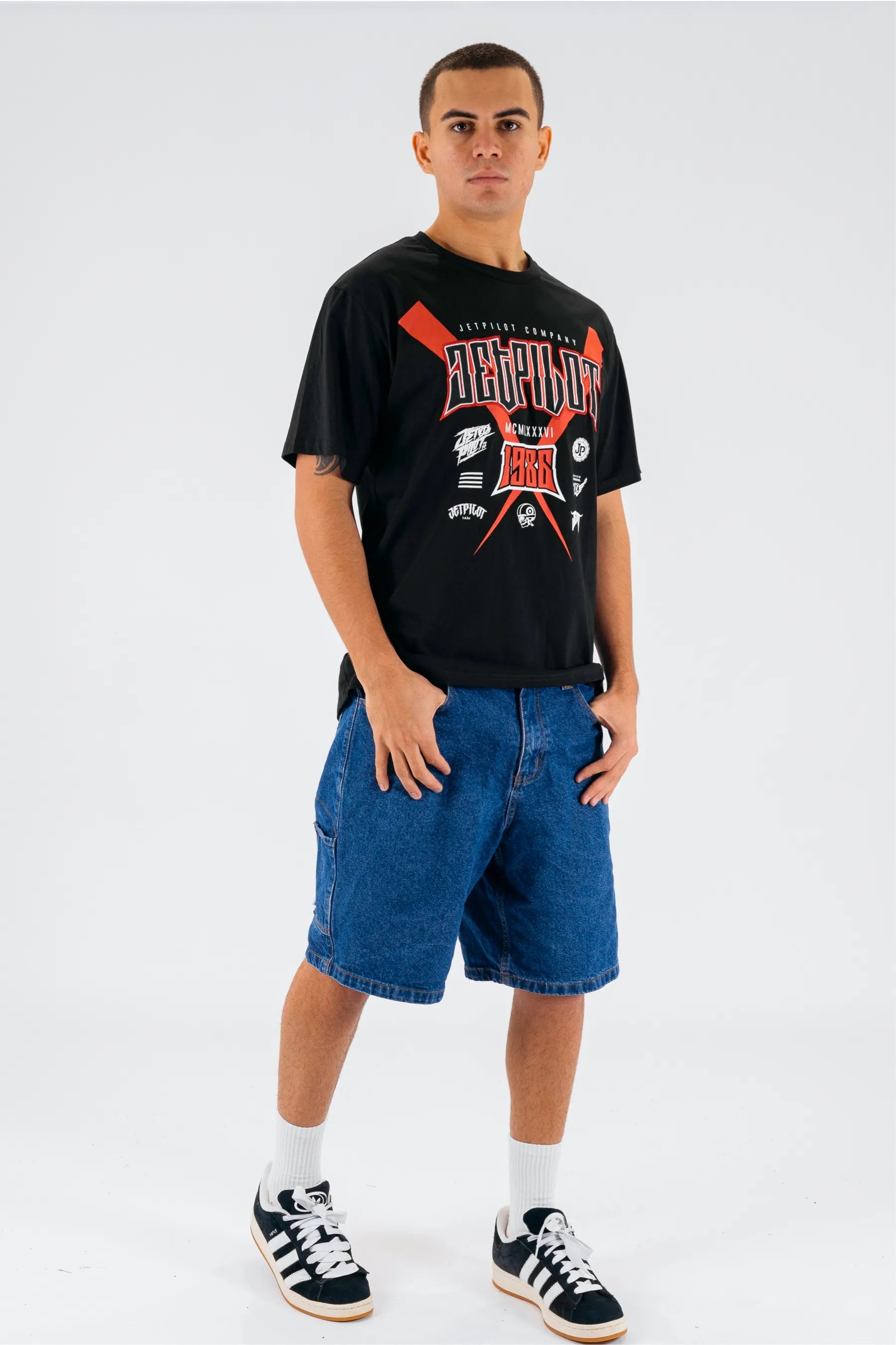Person wearing a black t-shirt with graphics and text, blue shorts, and black sneakers on a white background — view 4 (3)