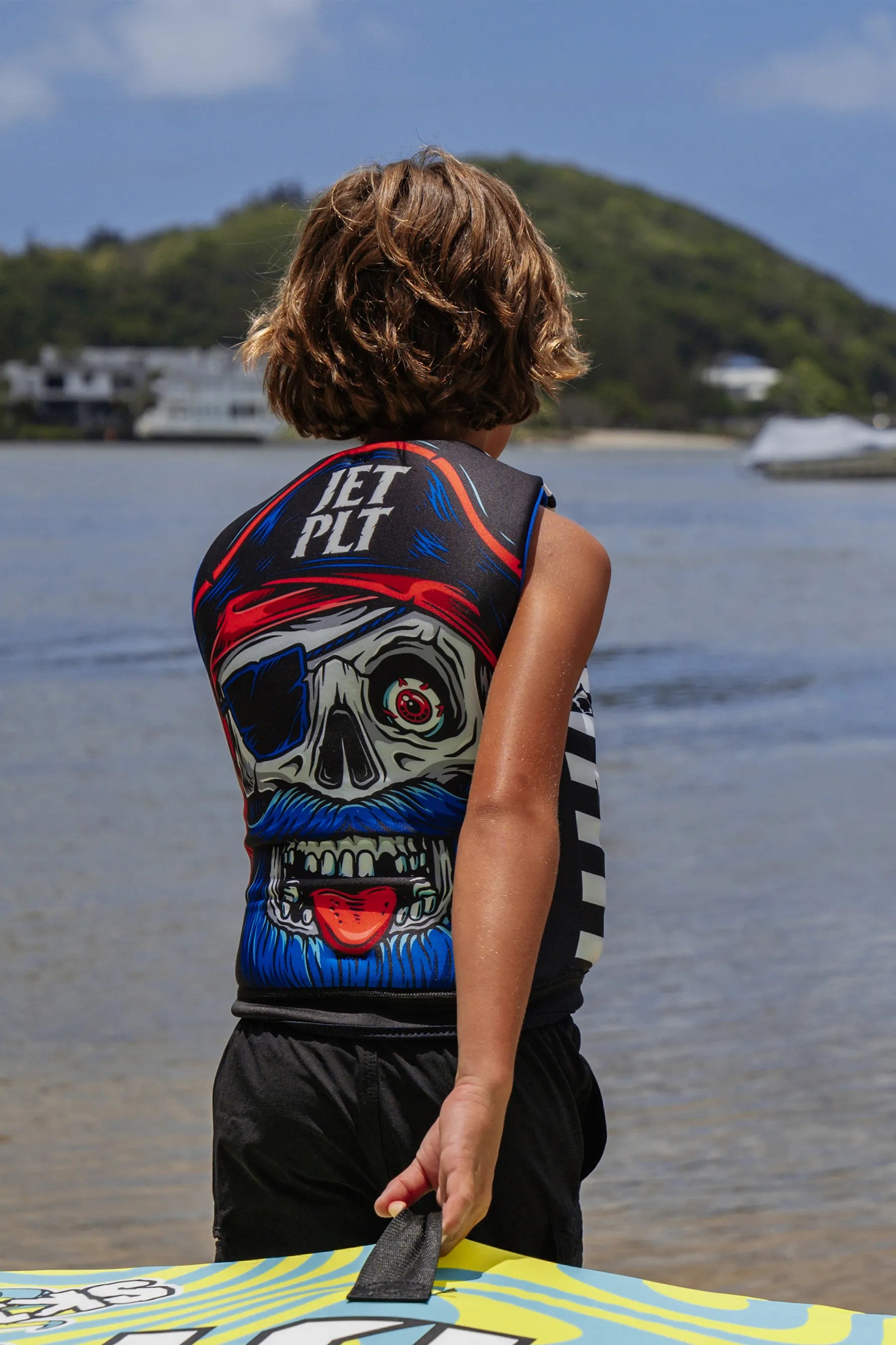 child wearing a colorful life jacket with a skull pirate design by a body of water.