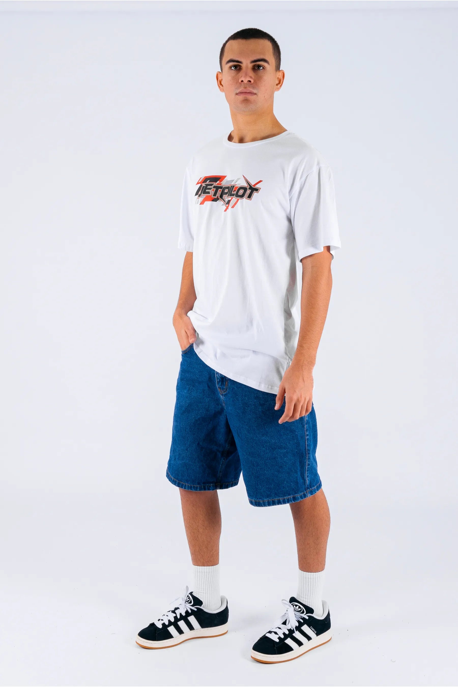 A person wearing a white tee with a Jetpilot logo, blue shorts and black/white on a white background — view 3 (3)