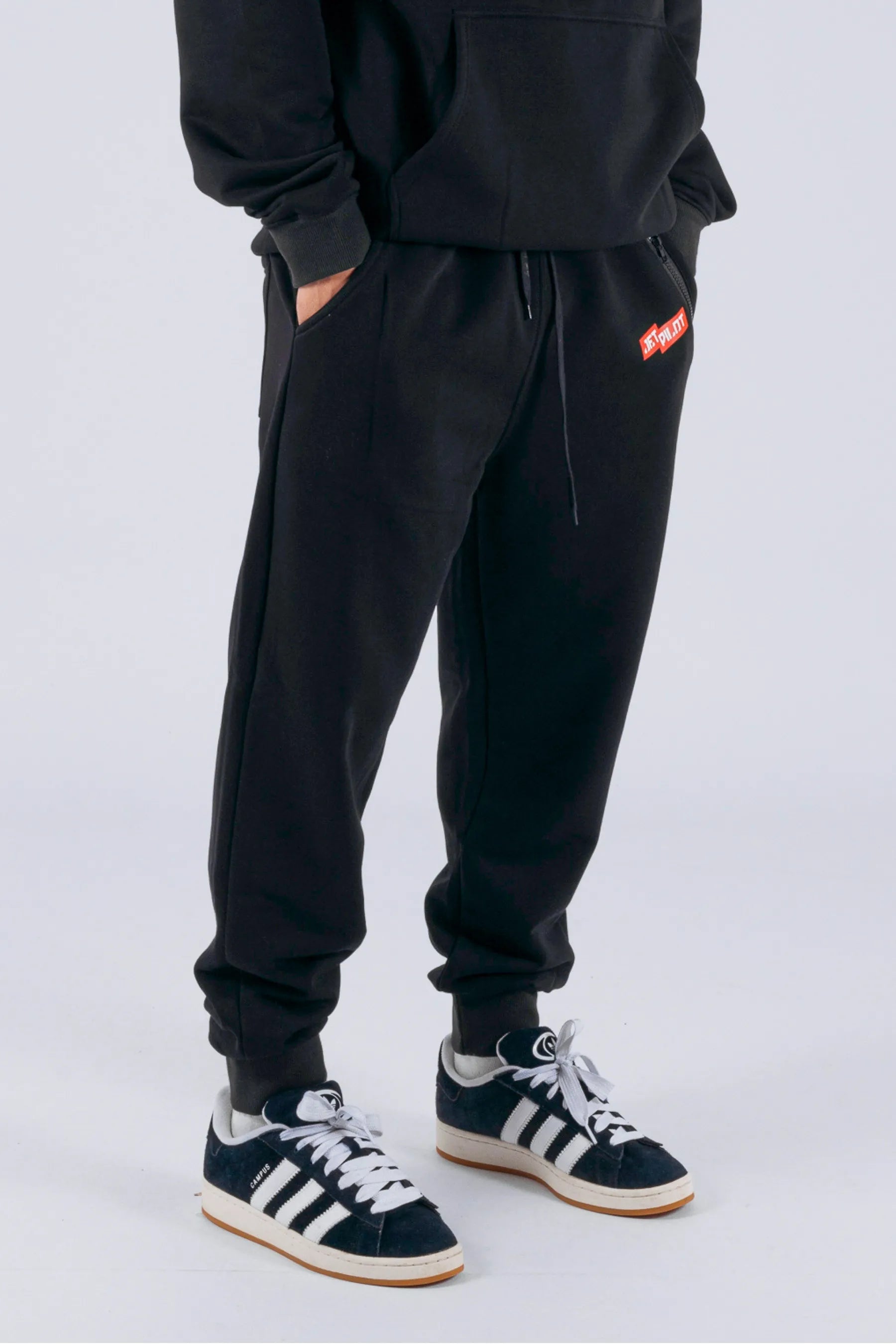 Right side view of a person wearing black trackpants with a red Jetpilot logo on a white background — view 3 (3)