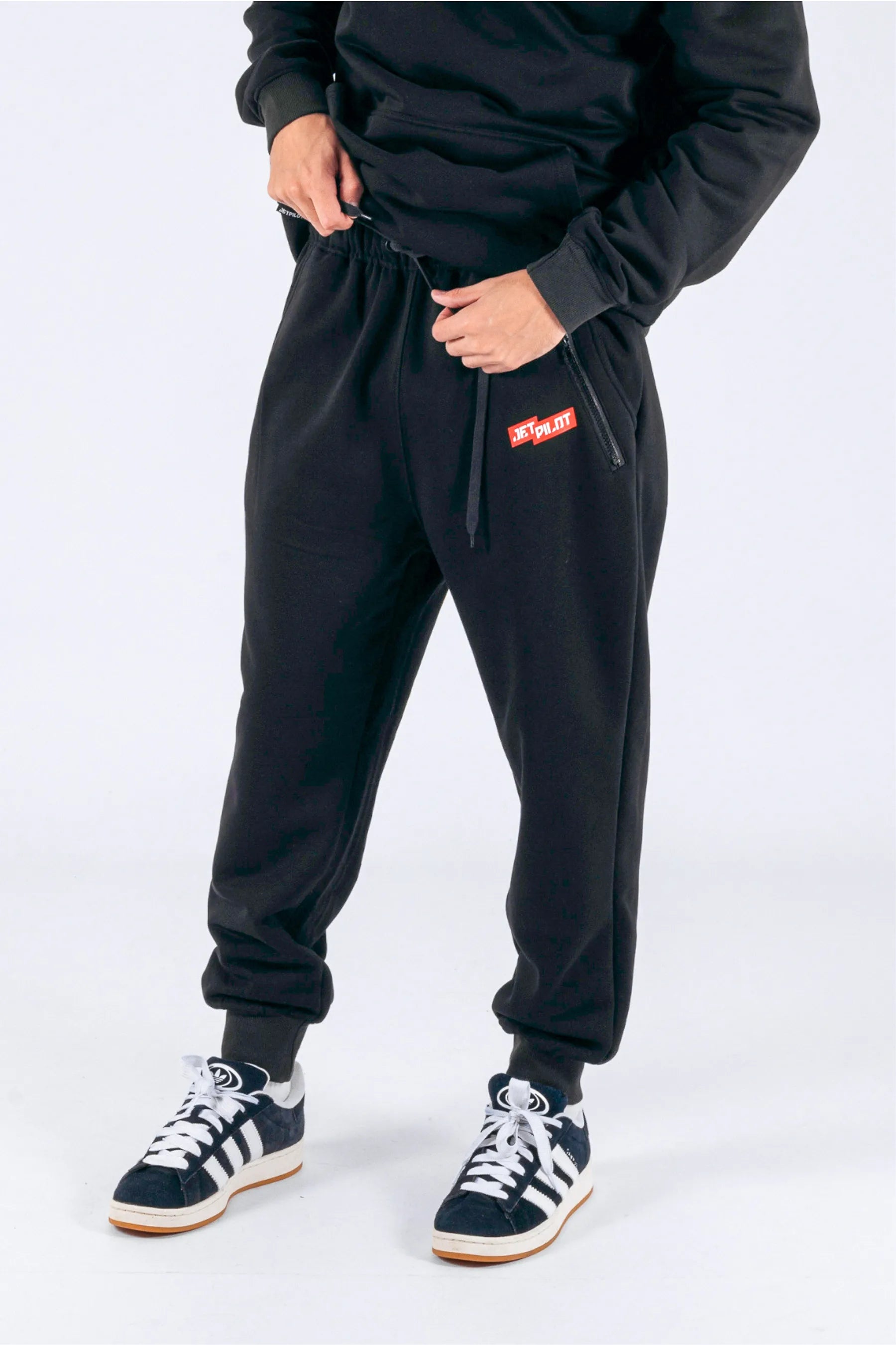 Left side view of a person wearing black trackpants with a red Jetpilot logo on a white background — view 4 (3)