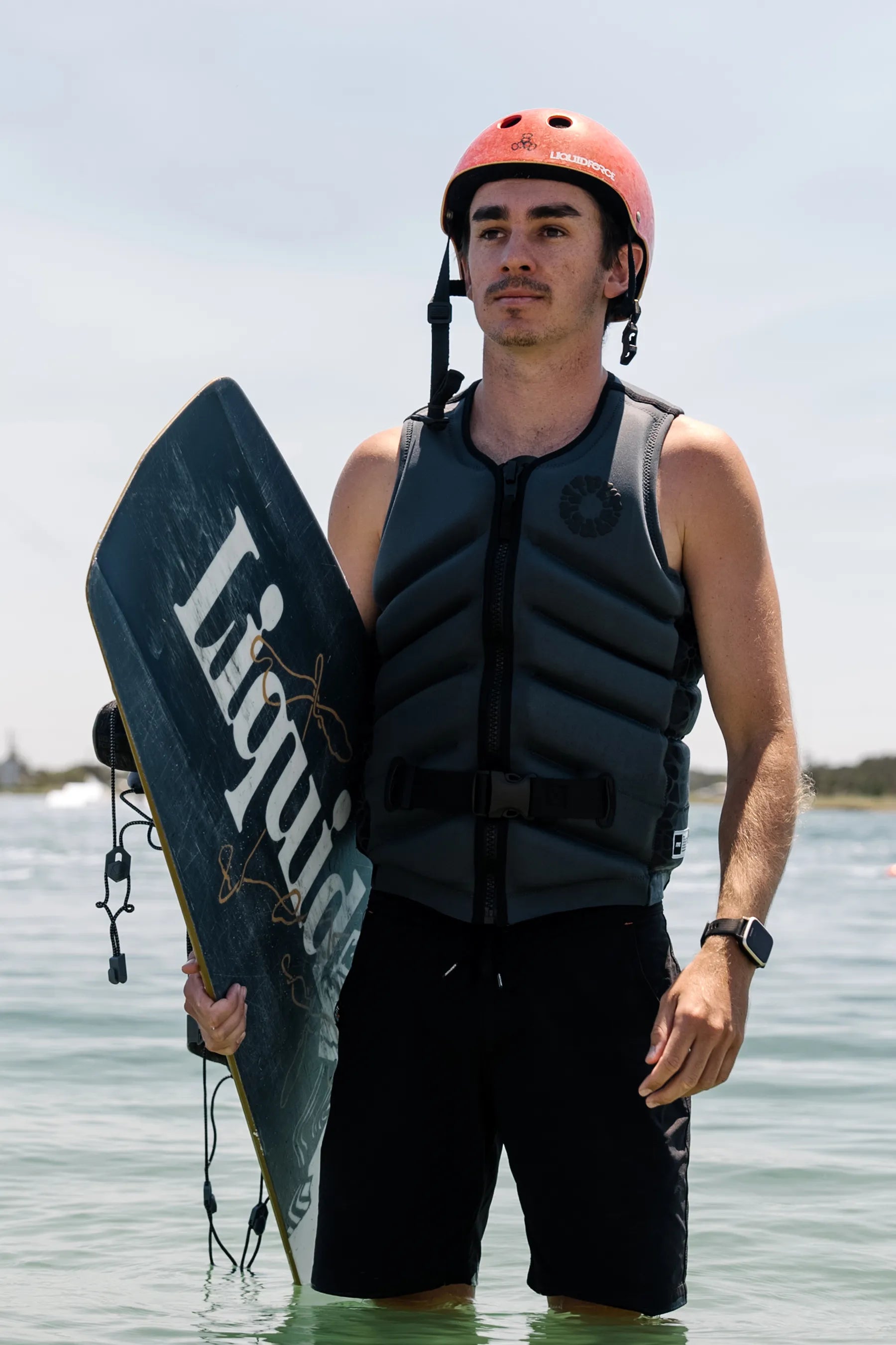 Person holding a wakeboard with a helmet and Jetpilot X1 Life Jacket - Bust Dunn Signature Series - Charcoal, standing in water.